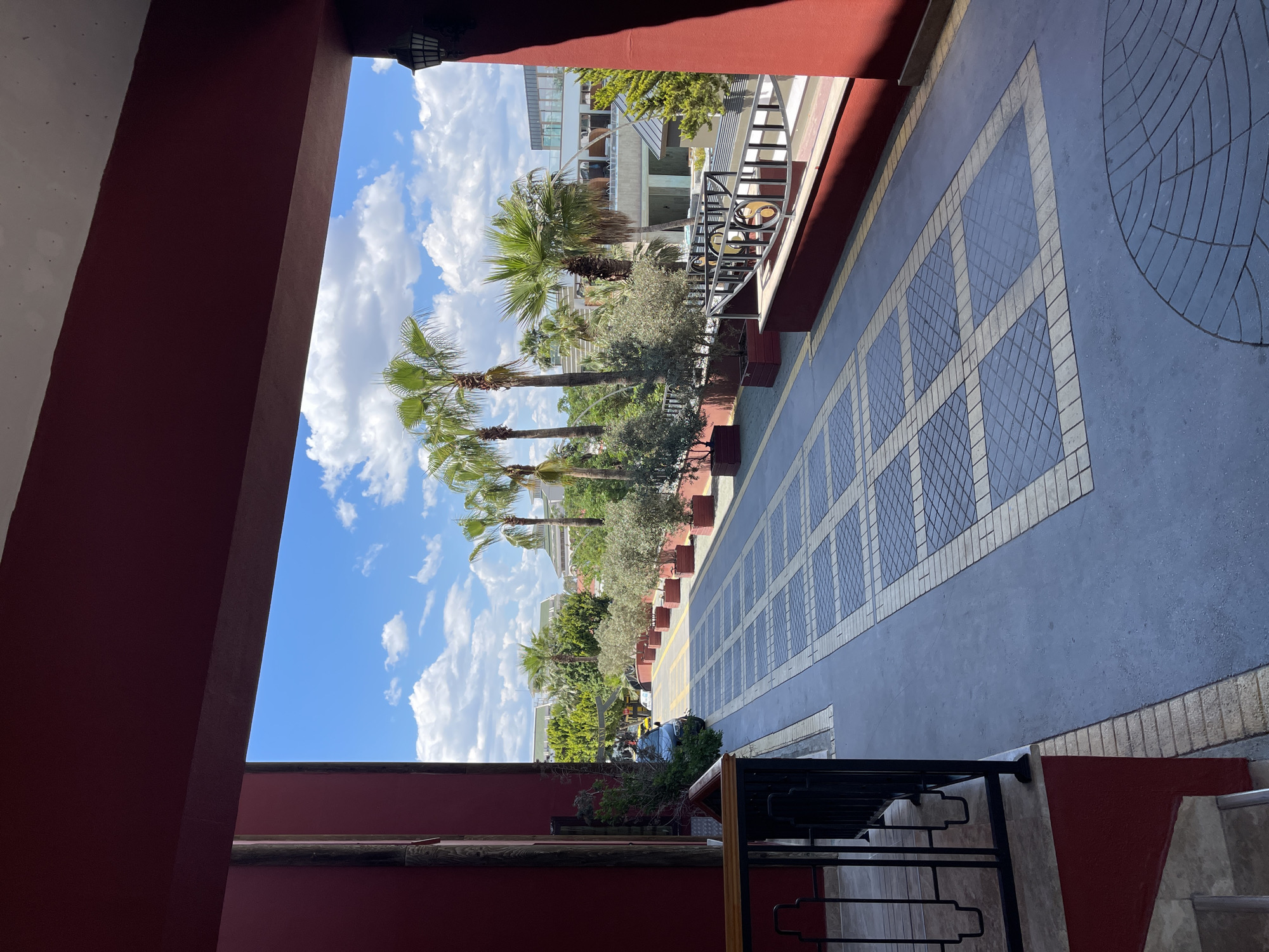View looking out from a building, possibly a hotel or resort, in an unidentified location. The frame is defined by red walls. Outside, a tiled walkway with a decorative pattern stretches into the distance. Palm trees and other greenery line the side of the walkway, partially obscuring a building beyond. The sky is blue with scattered white clouds. A black railing is visible in the foreground.