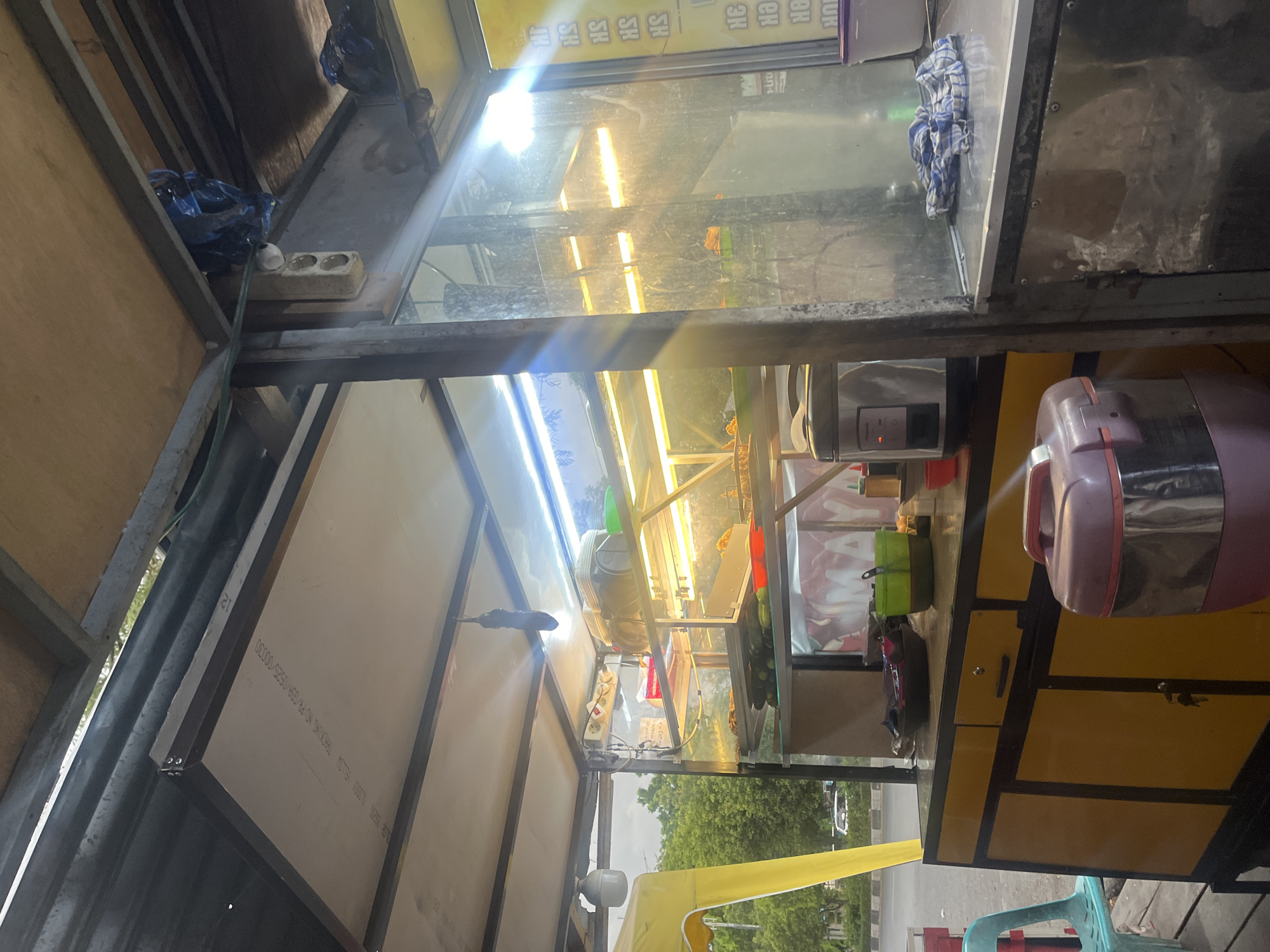 An outdoor food stall in Banda Aceh, Indonesia, features a display case filled with food. The stall has a metal frame and a yellow counter with drawers and cabinets. A rice cooker and various containers are visible on the counter. The interior of the stall is lit by warm, fluorescent lights, creating a glowing effect through the glass of the display case. Outside the stall, a yellow tent is partially visible, along with trees and a street with a car. The overall scene depicts a typical street food vendor setup in a tropical environment.