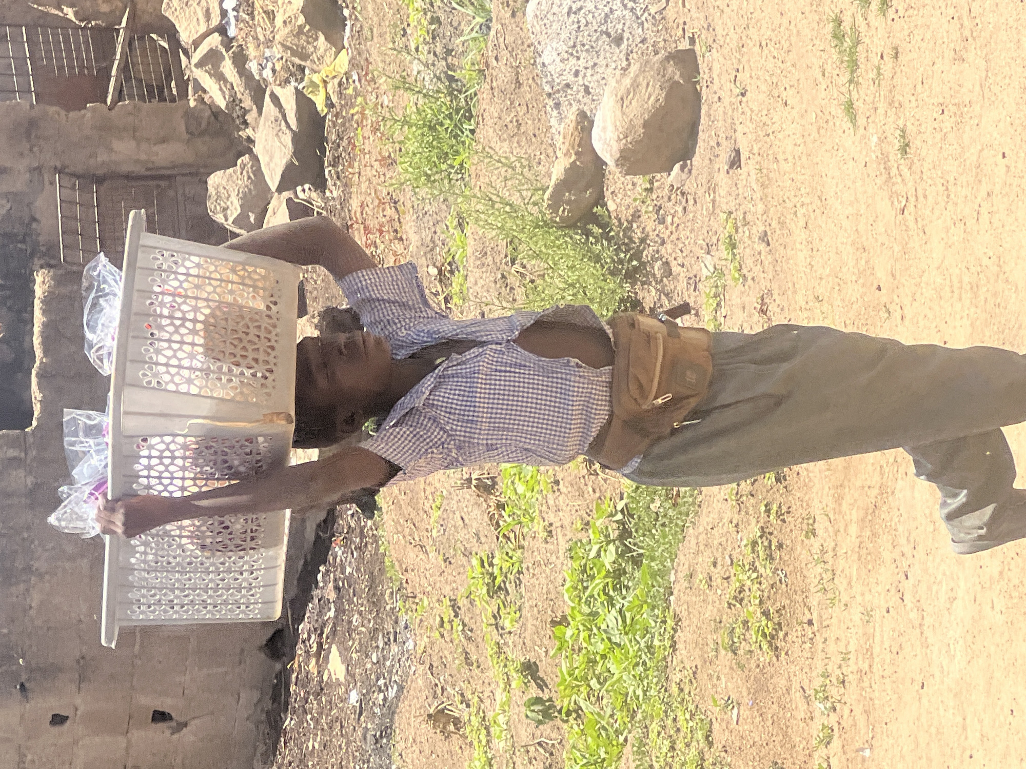 A man is walking on a dirt road in what is presumed to be the city of Gura Top, Nigeria. He is carrying a stack of two white plastic baskets filled with items wrapped in clear plastic overhead. The man is wearing a blue checkered short-sleeved shirt, gray pants, and has a small brown pouch or bag strapped around his waist. To his left is a dilapidated building of gray concrete or stone. To his right, the ground is dirt and overgrown with small weeds. In the background are more buildings of similar construction, and there are rocks scattered in the background.