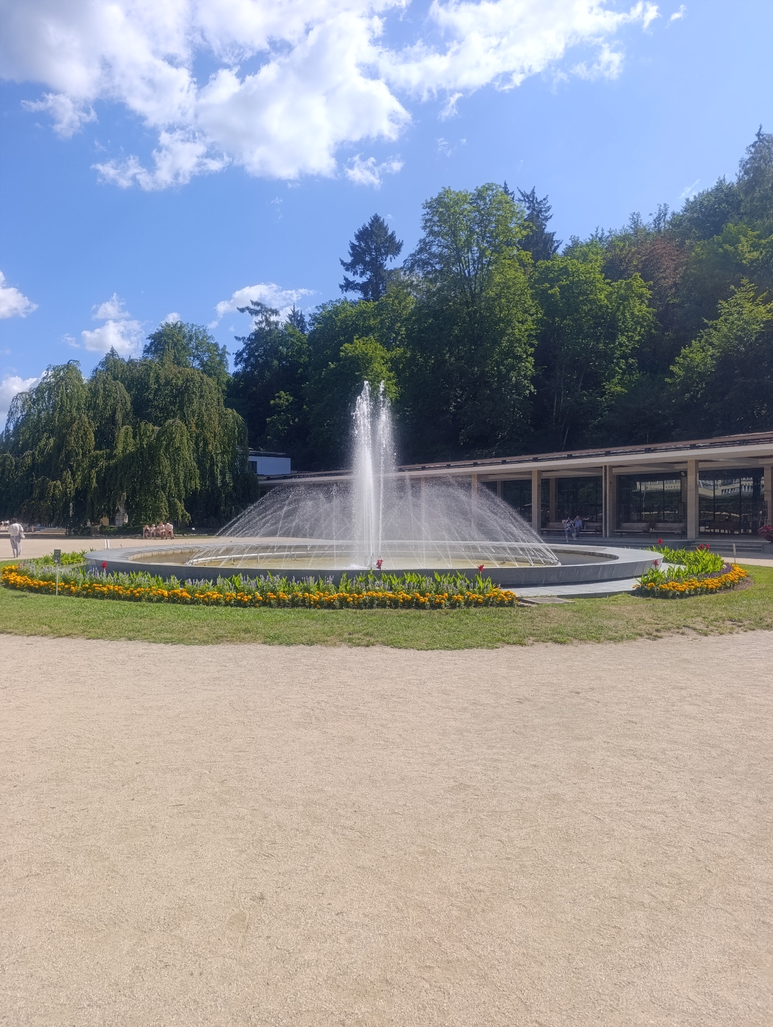 The image depicts a frontal view of a large, circular fountain in a park setting in Luhačovice, Czechia. The fountain is actively spraying water upwards and outwards, creating a misty effect. Surrounding the fountain is a manicured garden with rows of yellow and orange flowers, interspersed with green foliage. Beyond the fountain and gardens, a colonnade structure with a covered walkway and open-air seating areas is visible. Several people are seated on benches within the colonnade. In the background, a dense forest of green trees rises up a hillside, and the sky is blue with scattered white clouds. The foreground is dominated by a wide expanse of light brown, sandy ground. The overall scene suggests a tranquil and well-maintained public space.