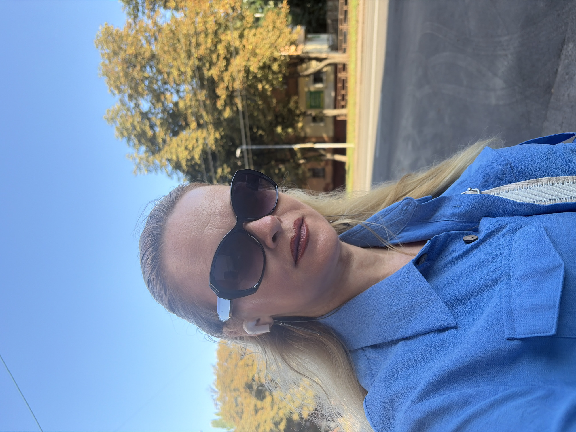 A woman with blonde hair, wearing sunglasses and a blue shirt, takes a selfie outdoors. She has on white earbuds. In the background, trees with yellow leaves and a building are visible, along with a street and a clear blue sky.