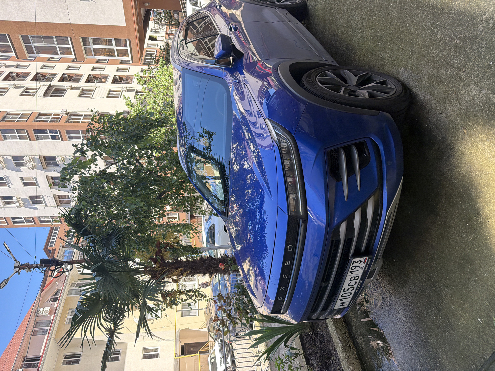This image depicts a sunny street scene in a residential area, consistent with the context of Sochi, Russia. The primary subject is a vibrant blue EXEED SUV, parked on the street with its front facing slightly left. The car's polished surface reflects the surrounding buildings and foliage.
The front license plate of the blue SUV is clearly visible, bearing the text "M105CB 193 RUSSIA." The "193" region code corresponds to Krasnodar Krai, where Sochi is located.
In the background, tall, multi-story residential buildings with light-colored facades and brown accents are prominent. These buildings feature numerous windows, many with external air conditioning units. Lush green trees, including a large palm tree and a deciduous tree, are interspersed throughout the scene, partially obscuring the buildings and casting reflections on the car. Overhead wires and a street lamp are also visible against the clear blue sky.
The ground appears to be asphalt or concrete, showing some reflective dampness. The scene is brightly lit by direct sunlight, indicating daytime. No individuals or specific activities are depicted, portraying a serene urban environment. A lighter-colored vehicle is partially visible parked behind the blue SUV.