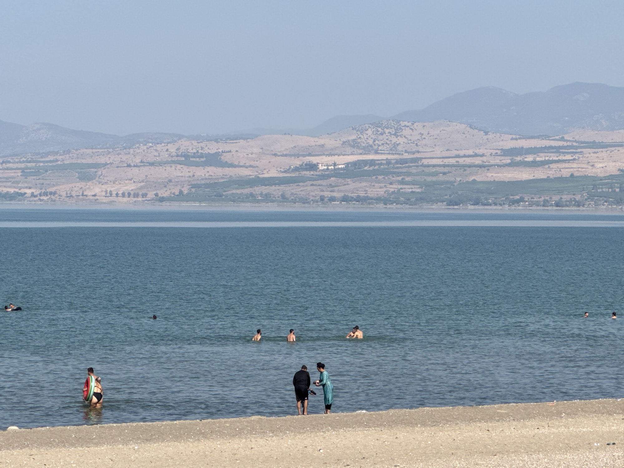 The image depicts a scenic view of the Sea of Galilee in the Golan Regional Council, Israel. The water is a calm, deep blue, stretching towards a hazy horizon where rolling, dry, tan-colored hills meet the sky. Patches of green vegetation dot the hillsides, and a few small structures are visible in the distance. The foreground features a sandy beach, sparsely populated.
In the water, several individuals are engaged in recreational activities. Two people are standing near the shore, one wearing a red top and black bottoms, holding a green inflatable ring, and the other is partially submerged. Further out, a group of men are swimming. One person is seen floating on their back further out to sea, and another is swimming. Closer to the shore, two figures, one in a dark outfit and another in a turquoise outfit, are standing on the wet sand. The sky is a pale, hazy blue, indicating a clear but possibly warm day. The overall atmosphere is one of a peaceful day at the beach.