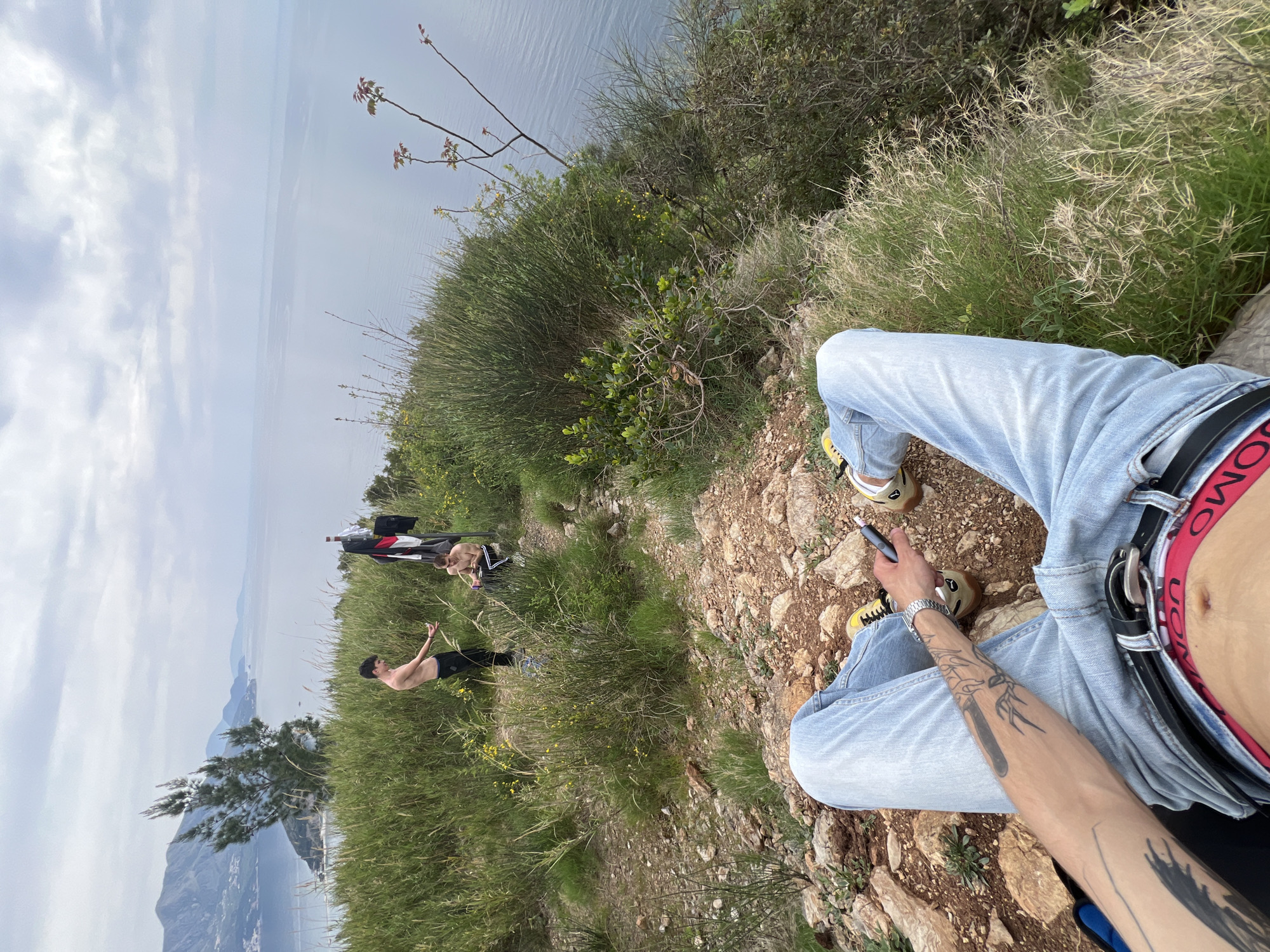 A first-person perspective of a person sitting on a rocky and grassy hillside, overlooking a body of water and mountainous terrain. The person, wearing light blue jeans, brown and yellow shoes, and a black belt with a red Uomo band, holds a vaping pen in their right hand. The person's arm is tattooed with various designs. Further along the hillside, two people are visible in the distance. One is shirtless and wearing black pants, gesturing with their arms, while the other is sitting and smoking. There is a black bag and articles of clothing lying nearby. The scene appears to be located in Budva, Montenegro, based on the landscape.