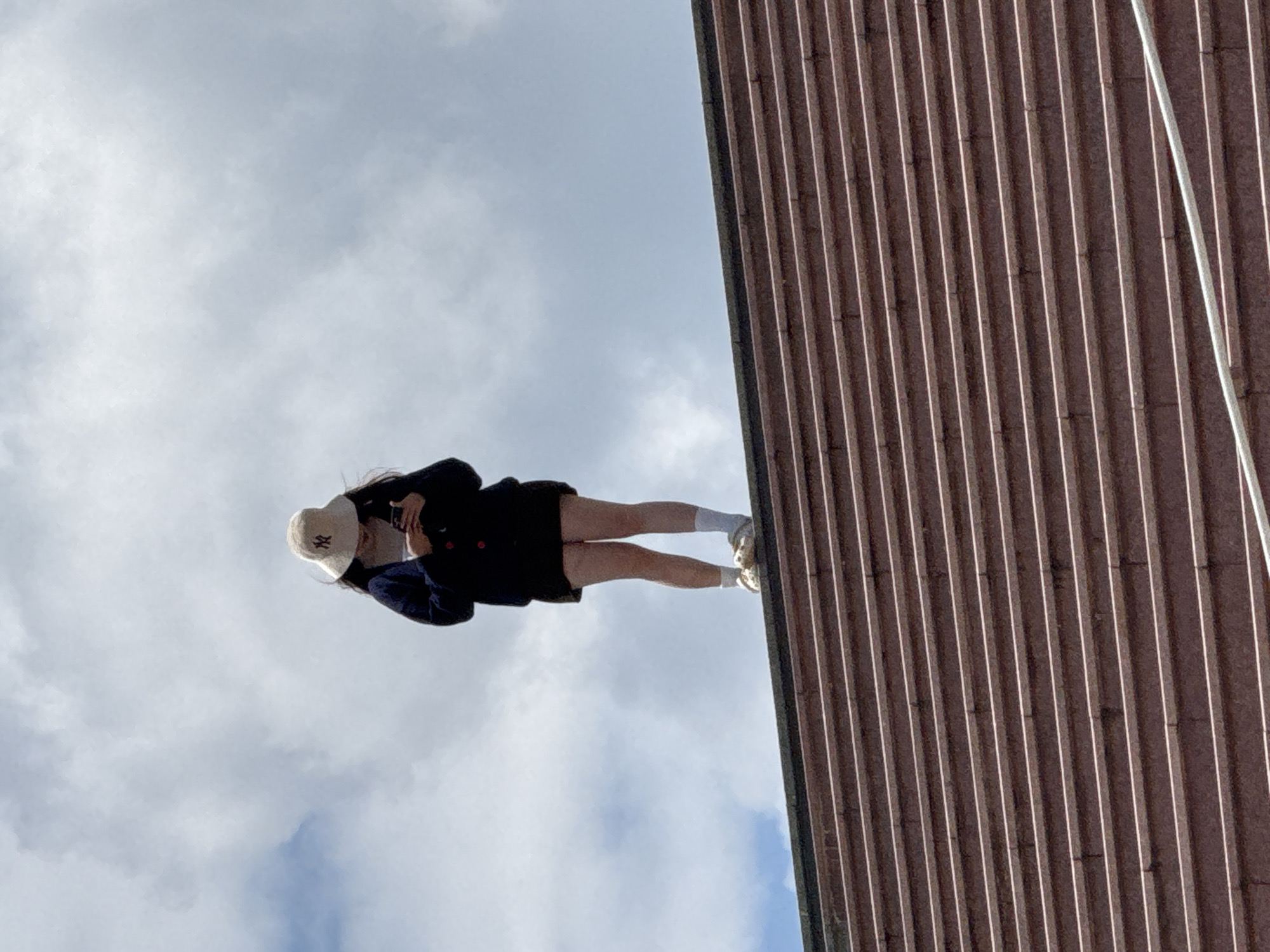 A lone individual, wearing a bucket hat and a dark sweater over a skirt, stands on the edge of a tiered structure, looking down at a mobile phone. The person is dressed casually in white socks and light-colored shoes. The tiered structure, possibly part of an outdoor amphitheater or grand staircase, is made of reddish-brown stone with light-colored strips. The background is dominated by a cloudy sky. The specific location in Da Lat, Vietnam, is not identifiable from the visual elements presented in the image.
