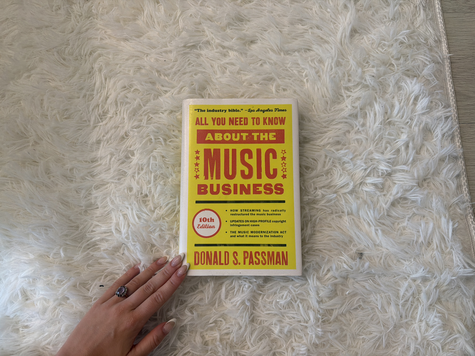 The image shows a copy of the book "All You Need to Know About the Music Business" by Donald S. Passman, 10th Edition, lying on a white, fluffy rug. A hand wearing a ring with a purple stone is also visible in the lower left corner. The rug appears to be on a wood floor. The image does not contain information about Seoul, South Korea.