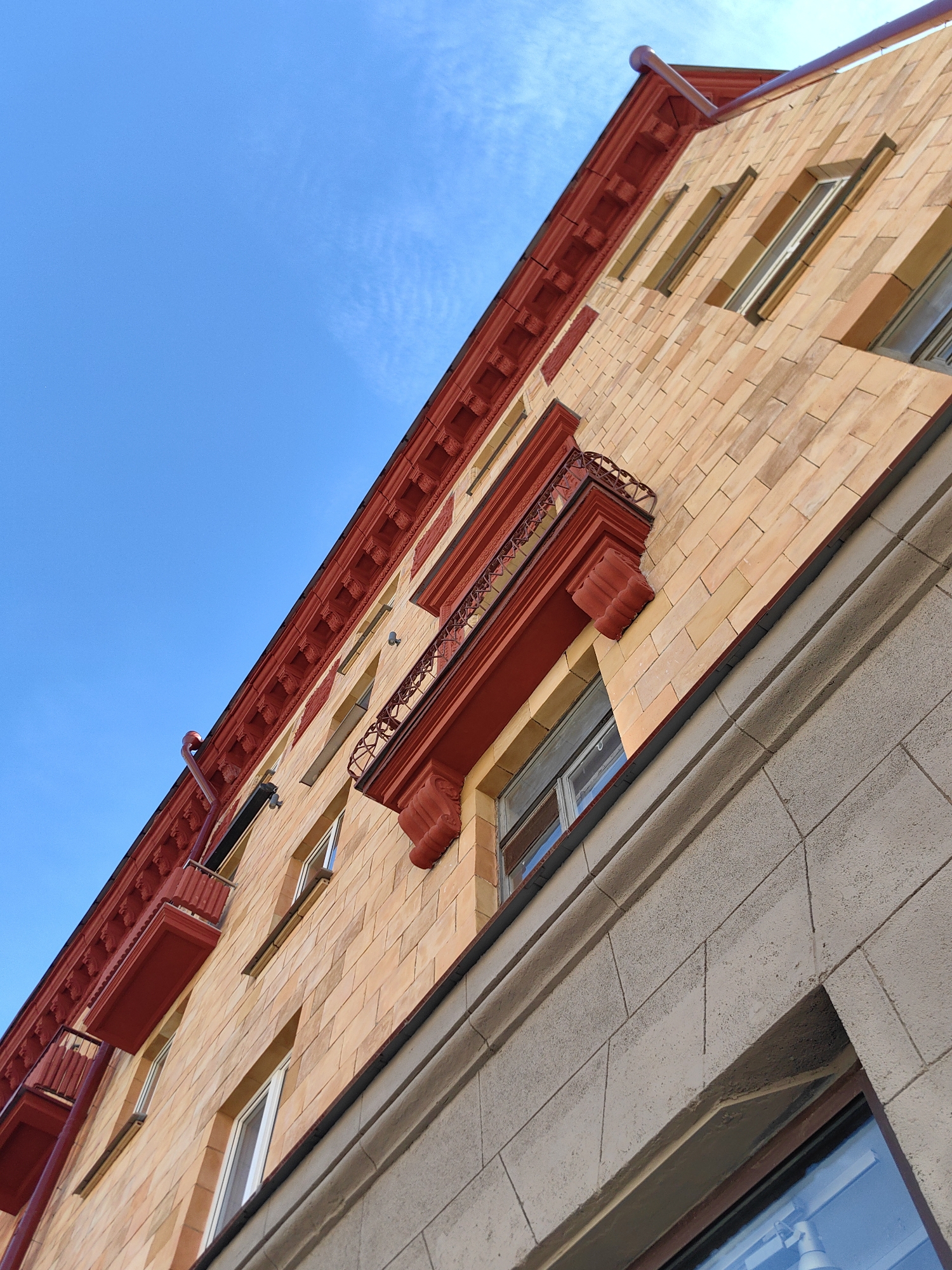 The image is a low-angle outdoor shot of a building's facade, predominantly featuring a clear blue sky with wispy clouds and a section of a building constructed with light orange brick. The building has distinctive red decorative elements, including ornate trim along the roofline, balconies with intricate ironwork, and window surrounds. Several windows are visible, some of which are open. The lower portion of the building's facade is clad in large, light gray stone blocks with visible mortar lines. The overall impression is of an older, architecturally detailed building. The context of Minsk, Belarus, suggests this is likely an urban setting. There are no people or animals visible in the image.