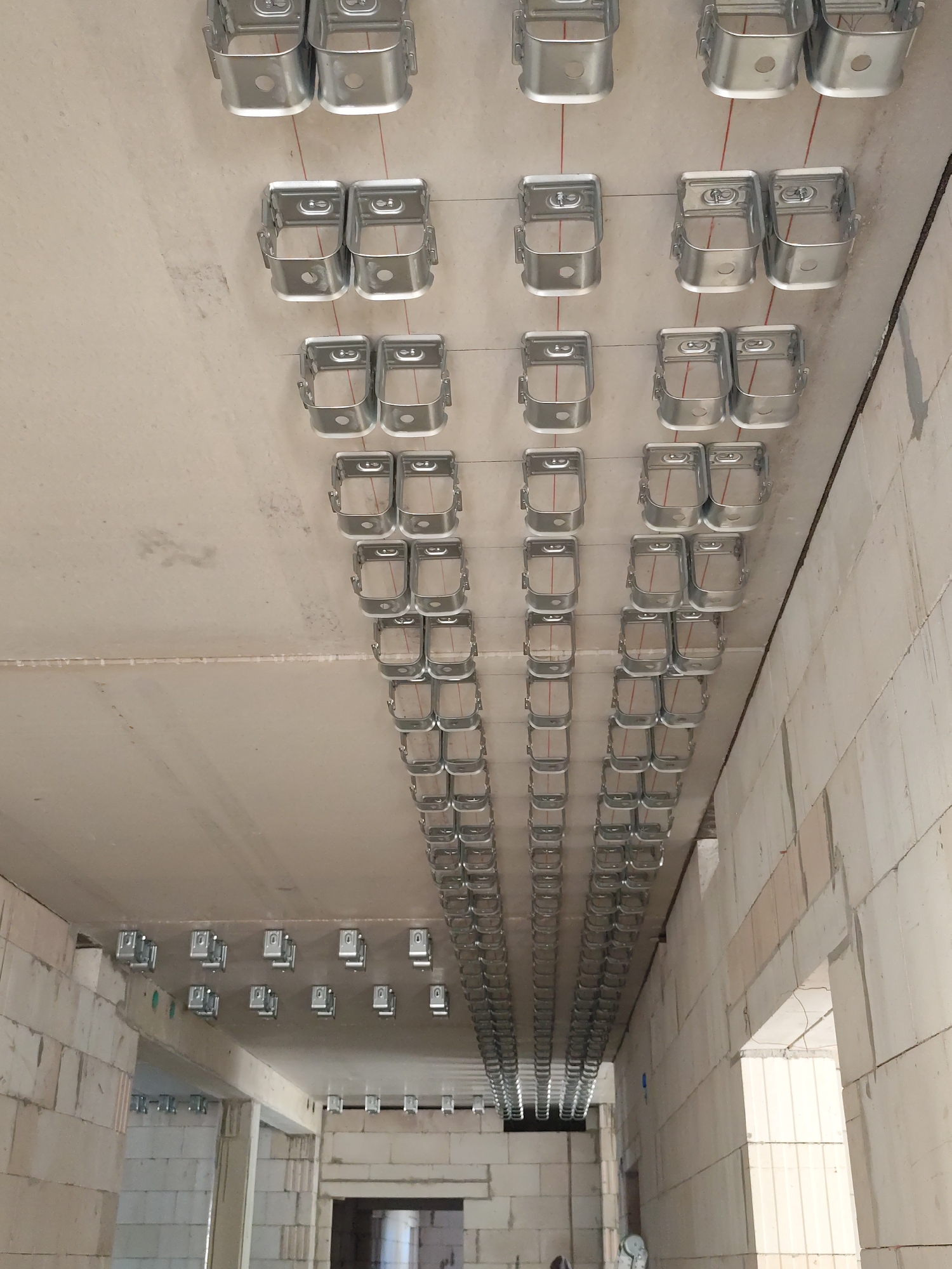 The image shows a ceiling in what appears to be a construction site. Numerous rows of metal brackets are attached to the concrete ceiling. These brackets are spaced evenly and are connected by thin red wires, suggesting they are part of a suspension system for lighting, electrical conduits, or other building services. The overall environment is unfinished, with exposed concrete and block walls visible in the background. There are no people or animals depicted in the image. The location is identified as Osterwieck, Germany, likely a building under construction or renovation.