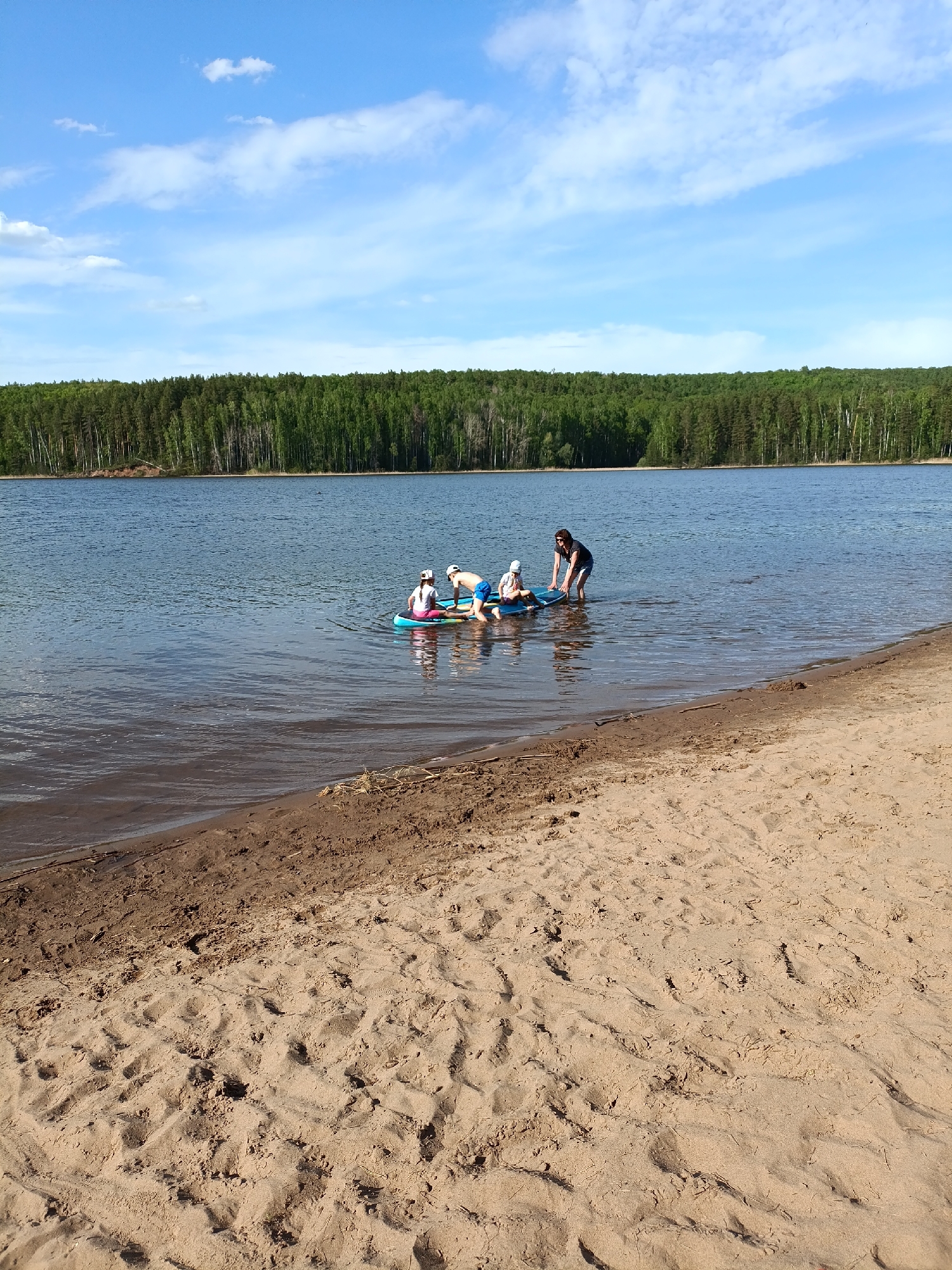 The image depicts a tranquil lake scene on a sunny day. A sandy beach occupies the foreground, gradually sloping into the water. In the middle ground, a blue inflatable paddleboard is afloat, carrying three individuals and a dog. One person is standing in the water, assisting the board. The background showcases a dense forest lining the opposite shore. Above, the sky is blue with scattered white clouds. The location is near Chaykovsky, Russia.