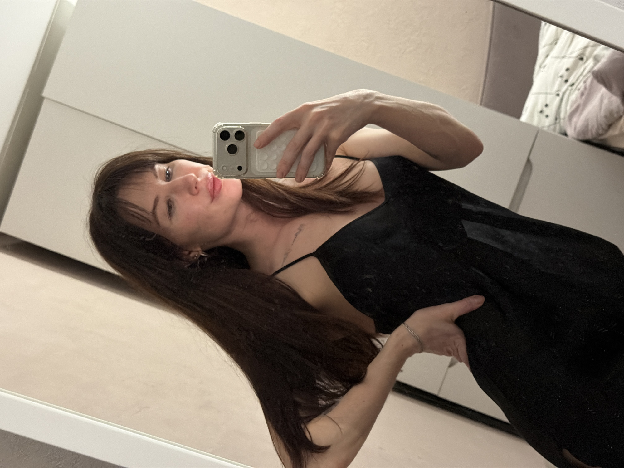 A woman is taking a selfie in a mirror. She has long brown hair with bangs, wearing a black dress with spaghetti straps. The woman is holding her phone in her right hand, positioned near her face. The background reveals a wardrobe and a glimpse of a bed with patterned bedding, suggesting the location is indoors, likely a bedroom. The city of Perm, Russia, cannot be confirmed from the provided image.