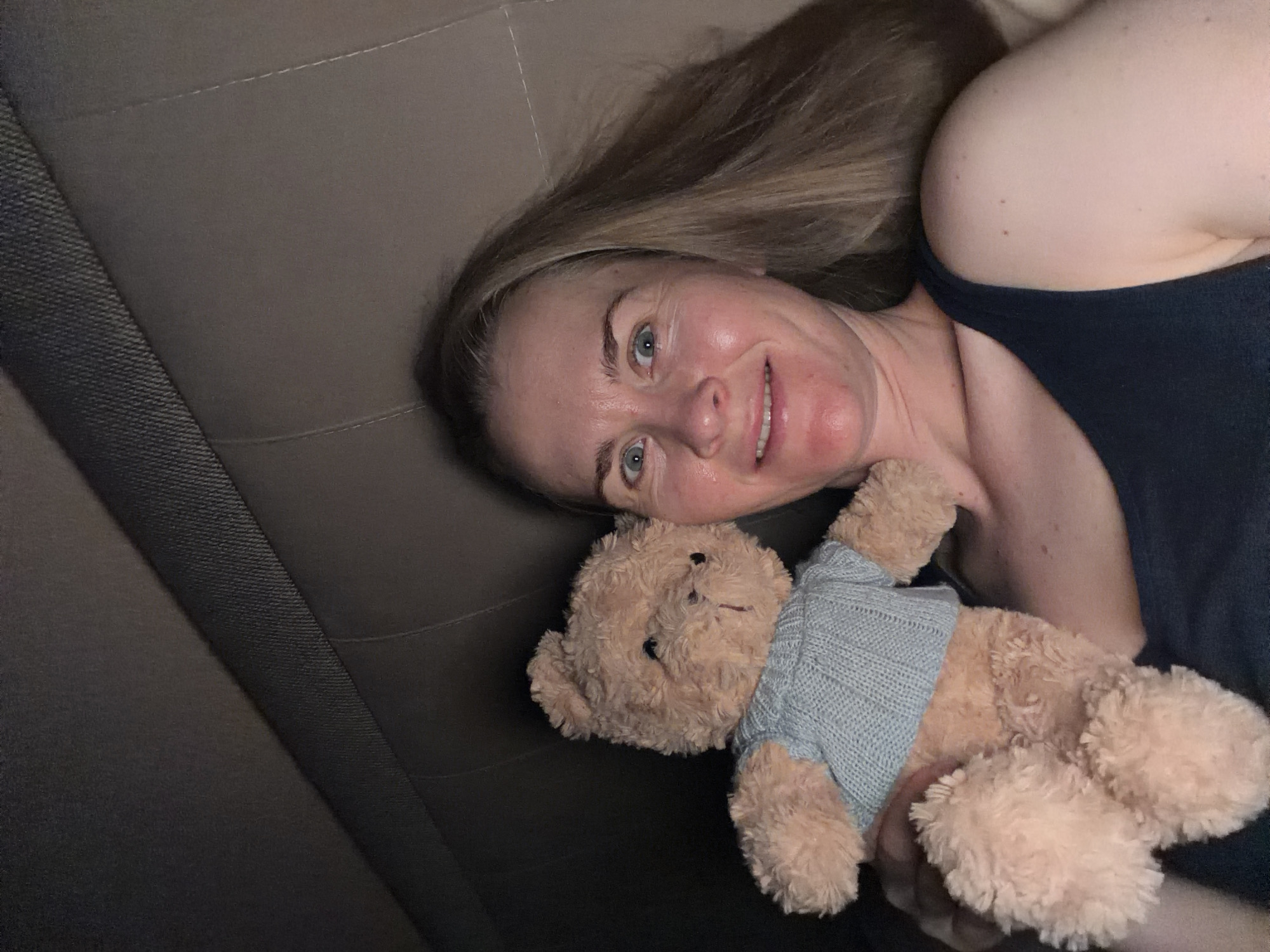 A woman with blonde hair and blue eyes is holding a light brown teddy bear wearing a blue knitted sweater. She is lying down, with the teddy bear held in her left arm. The background appears to be the interior of a vehicle, possibly a car, with a textured headrest. The woman is smiling and looking at the camera. The lighting is dim. The image was taken in Khabarovsk, Russia.