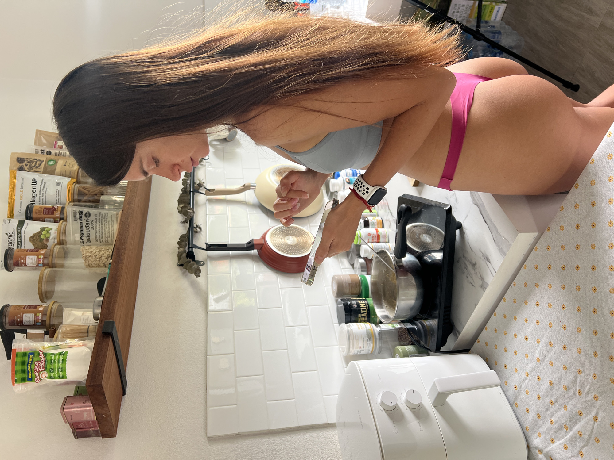 The image shows a woman in a kitchen, standing next to an air fryer and a stovetop where a pot is cooking. She is wearing a grey sports bra and pink underwear, and she is looking at her phone. On a shelf above the counter, there are various food items and containers. The kitchen appears to be a home setting. The image does not contain any information that would allow for the determination of its location in the city of บ้านบางรักษ์, Thailand.