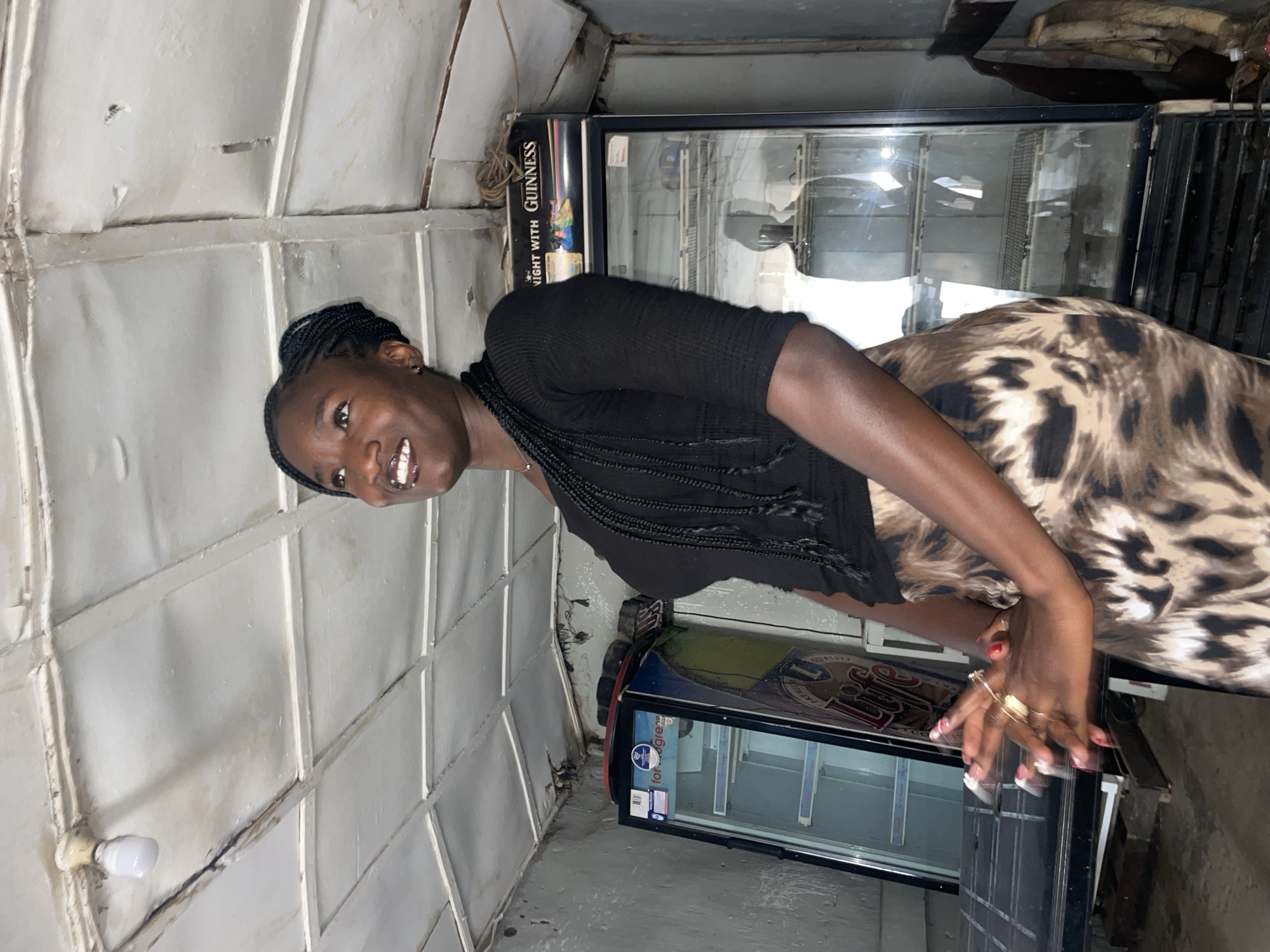 A medium shot captures a young woman standing in what appears to be an indoor commercial space, likely a shop or bar, in Jos, Nigeria. The woman, of African descent, has dark skin and is smiling broadly, looking directly at the camera. Her hair is styled in dark braids. She wears a black, short-sleeved top and a brown and black leopard-print skirt. Her hands are visible, showing manicured nails with white and red tips, and several rings on her fingers.
Behind her are two upright display refrigerators, typical of beverage coolers. The top refrigerator has "NIGHT WITH GUINNESS" prominently displayed on its side panel. The lower refrigerator shows glimpses of text like "QUALITY" and "PREMIUM," along with a white label that reads "for grea". The walls and ceiling of the space are constructed from what looks like white-painted corrugated metal or paneling, giving the area a utilitarian or possibly makeshift appearance. A simple lightbulb fixture is visible on the wall/ceiling to the left. The floor is concrete. The overall impression is a casual, friendly portrait taken within a local business environment.