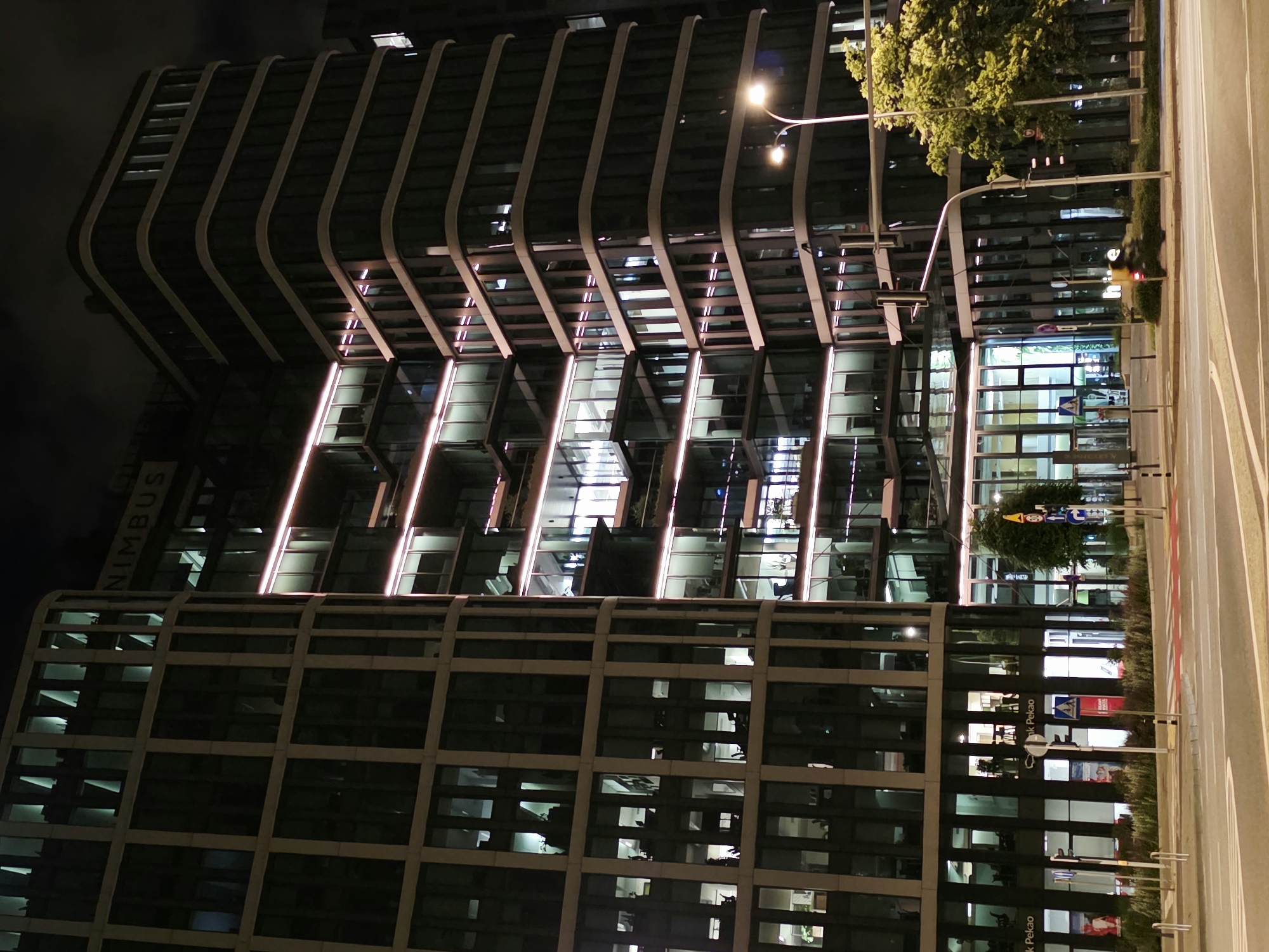 The image shows a modern office building at night in Warsaw, Poland. The building is illuminated with white and pink lights, highlighting its architectural features. The exterior is made of glass and concrete, with many windows revealing office spaces within. On the left side of the building, text that reads "NIMBUS" is visible. The building is situated on a street with a pedestrian crossing and traffic lights. A street lamp illuminates part of the scene. Trees and greenery are present on the right side, contrasting with the urban environment. The sky is dark, indicating it is nighttime.