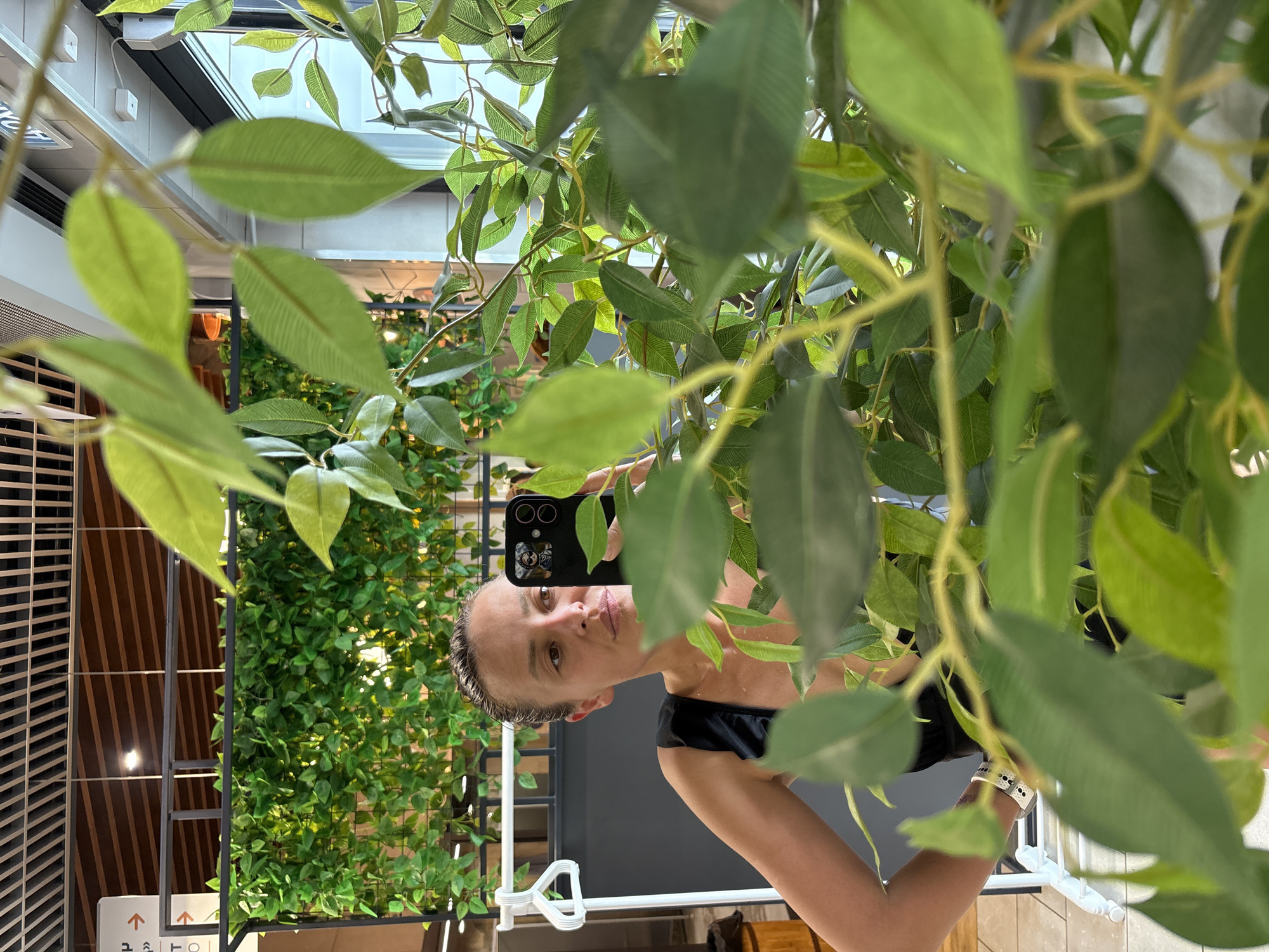 A person holds a smartphone aloft amidst a dense array of artificial green foliage, taking a selfie or photograph in Votkinsk, Russia. The subject, a fair-skinned individual, is partially obscured by the leaves, with the smartphone held at eye-level. The scene is composed of indoor foliage arrangements, visible behind and around the individual, in what appears to be a public space. The environment includes elements such as a ceiling structure and wall fixtures visible in the background.