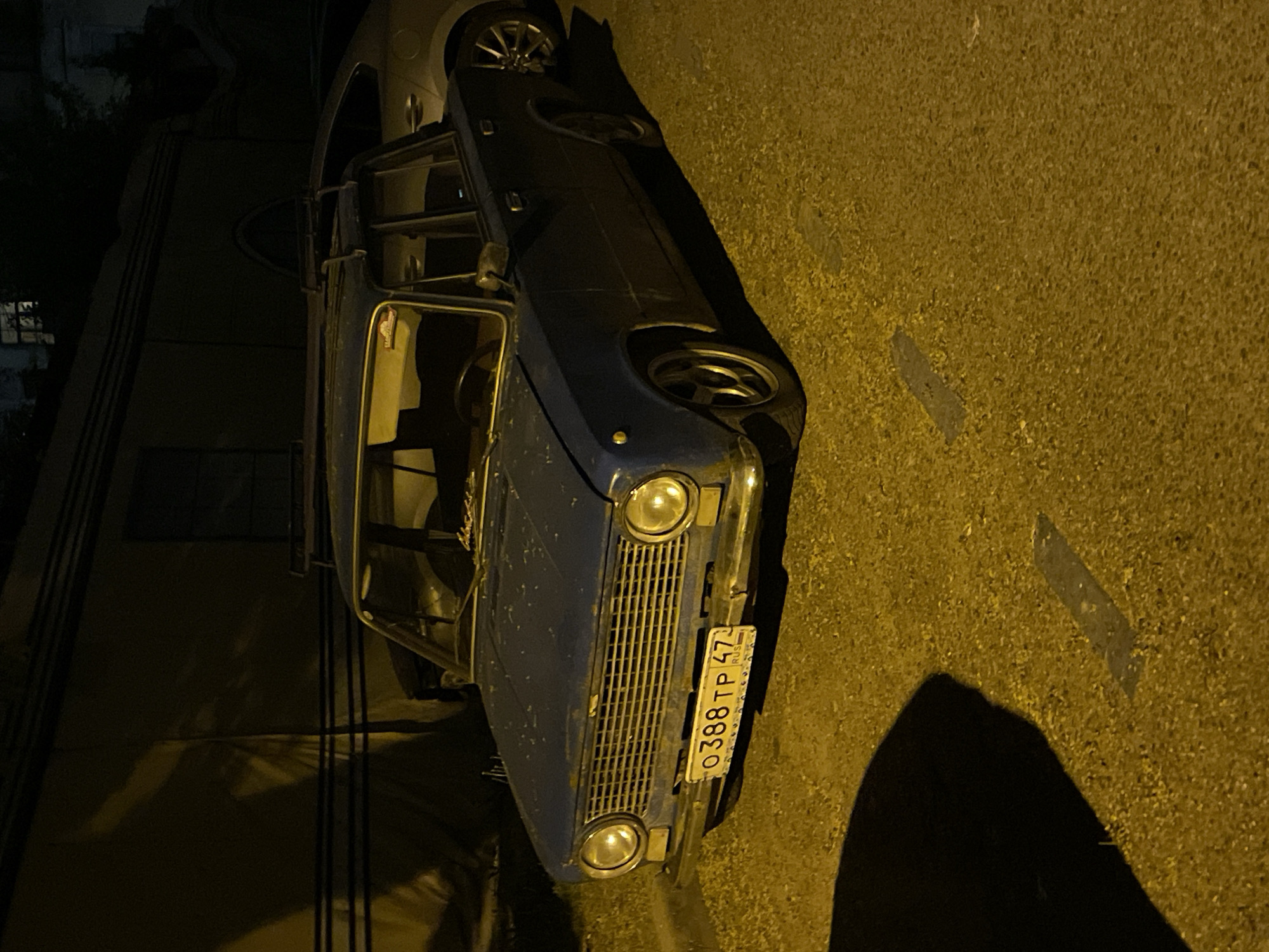 A dark blue Lada car is parked on a street in Saint Petersburg, Russia. The license plate reads "O388 TP 47 RUS". The car is parked next to a building. The street is dimly lit.