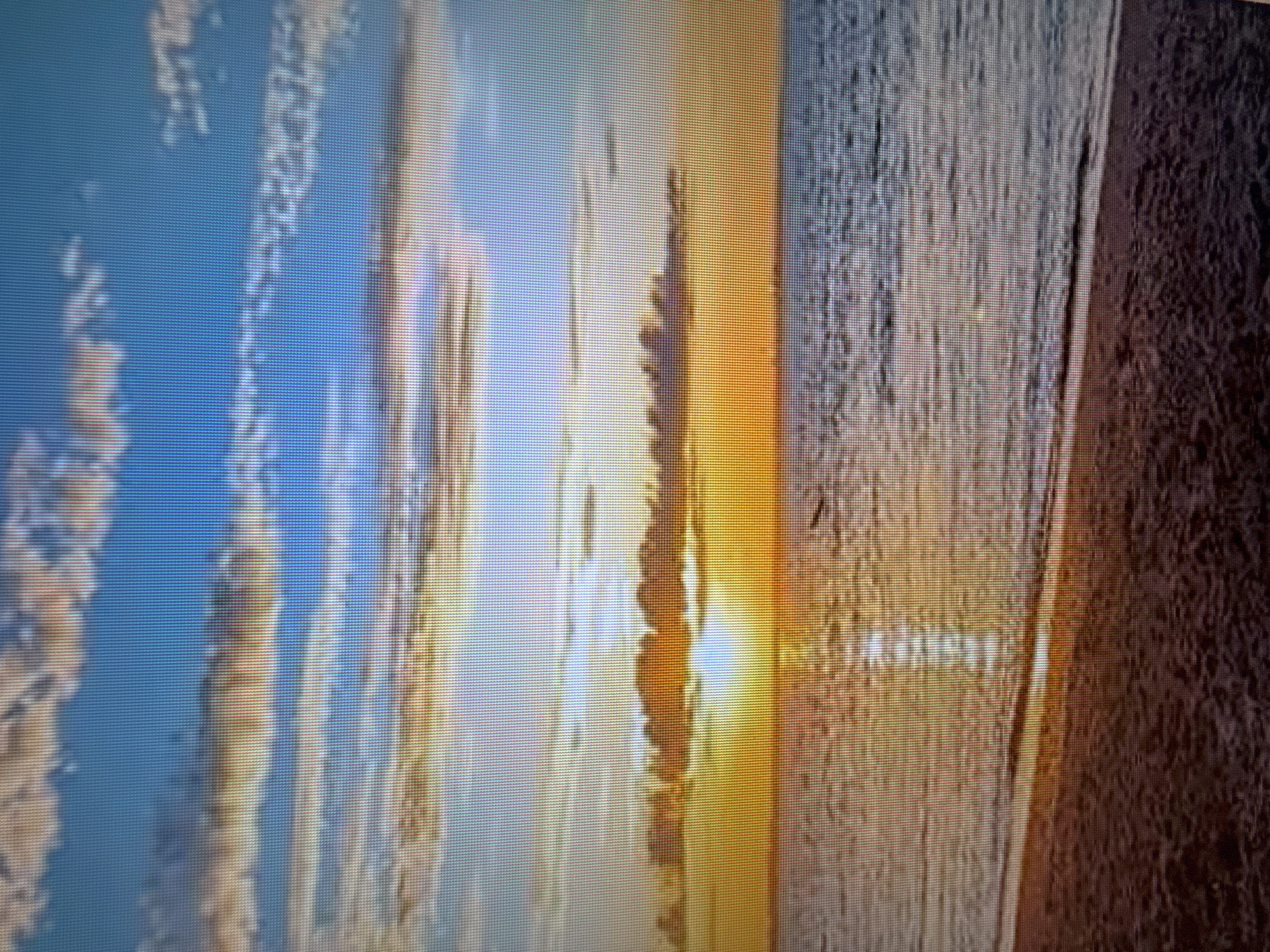 The image depicts a scenic view of a sunset over a body of water, likely the sea or a large lake, as suggested by the grainy texture of the water and the dark, pebbly shore on the right side of the frame. The sky is a vibrant mix of blues and oranges, with streaks of clouds illuminated by the setting sun. The sun itself is visible as a bright, horizontal glow near the horizon, casting a warm, golden light across the water and the sky. The overall impression is one of natural beauty and tranquility. There is no visible text in the image. The scene suggests a coastal or lakeside location during the evening hours.