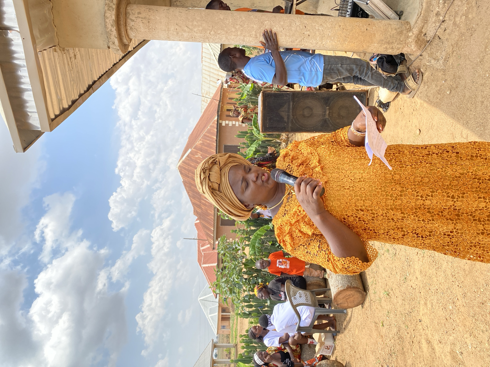 A woman in a gold lace dress and head wrap speaks into a microphone, holding a paper in her left hand. Behind her, a man in a blue shirt stands with his hands on a stone pillar, next to a large black speaker. In the background, a building with a red roof is visible, surrounded by trees and other structures. People are seated in the foreground, and the scene takes place outdoors on a dirt ground in Kuru Karama, Nigeria.