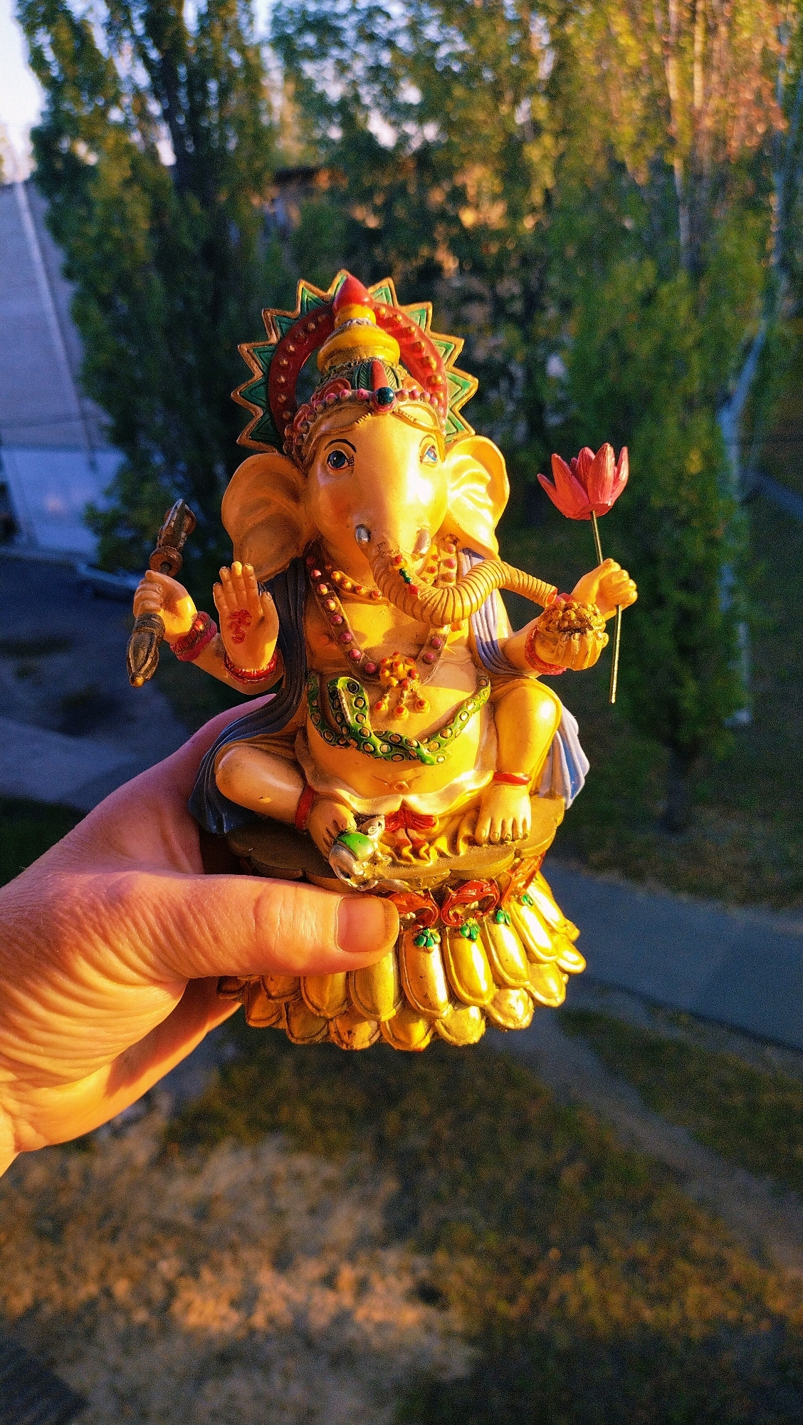 A hand holds a gilded statue of Ganesha, a Hindu deity with an elephant head. The statue is elaborately decorated with colorful details and holds a lotus flower. The background shows greenery and a blurred cityscape, possibly in Horishni Plavni, Ukraine, with the golden hour sunlight casting a warm glow.