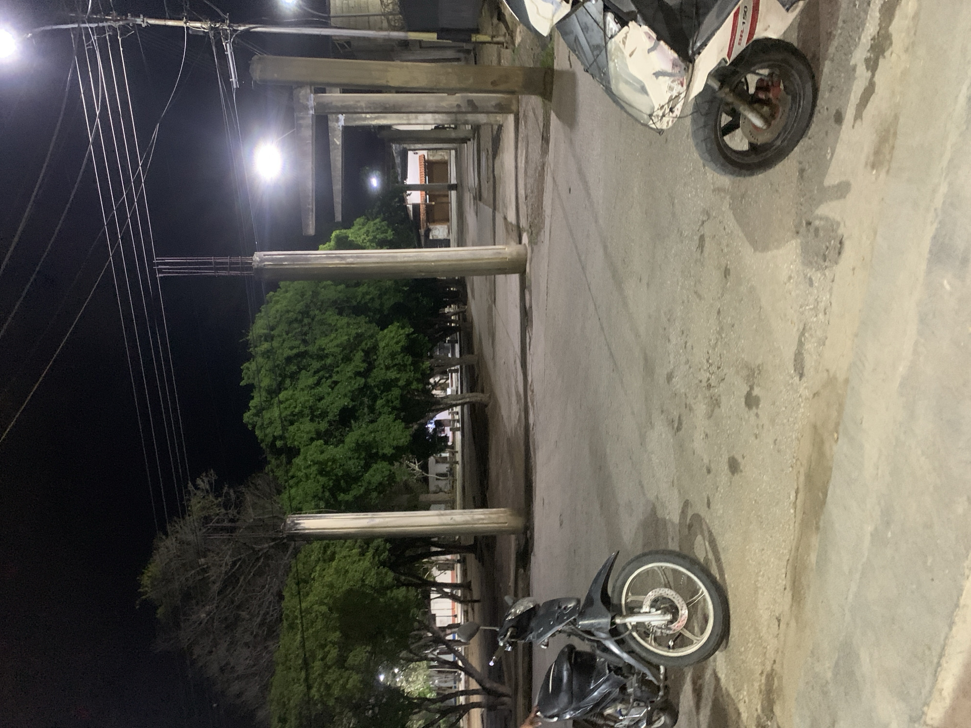 This night scene in Santa Ana, Venezuela, features a paved street or walkway flanked by a series of concrete pillars and trees. Two motorcycles are parked on the right side of the path. The closer motorcycle is dark-colored, likely black or dark grey, with its front wheel, disc brake, and engine partially visible. Further along, a white motorcycle with red accents is parked, displaying the text "BR-150" on its side fairing.
To the left, a row of substantial concrete pillars extends into the background, creating a colonnade that frames a view of structures or buildings further in the distance. Behind these pillars, a mix of dense, green-leafed trees and trees with bare branches are visible against the dark sky. Bright streetlights illuminate the area, casting light on the ground and reflecting off surfaces, with distinct lens flares from some of the light sources. Overhead on the far left, a complex network of power lines is silhouetted against the night sky. The ground shows some irregularities and stains, indicative of wear. The scene is devoid of people or specific activities, suggesting a quiet evening in an urban or semi-urban setting.