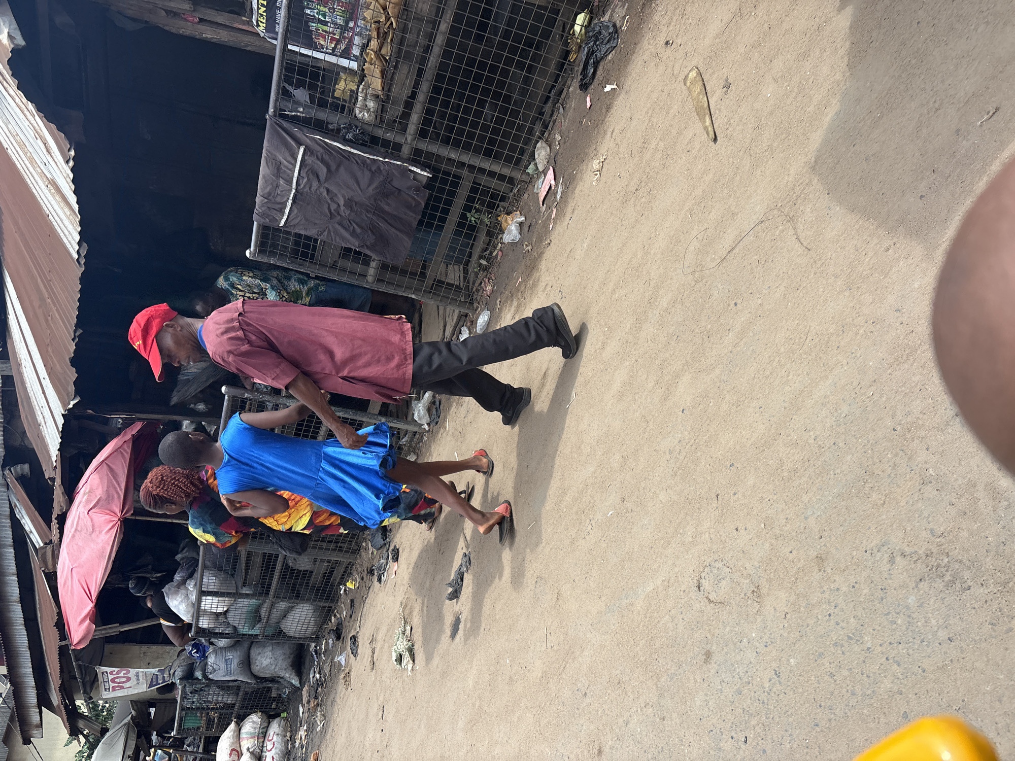 In the city of Fegge, Nigeria, a street scene unfolds under a weathered, corrugated metal roof of a market stall. A man in a maroon tunic and black trousers, wearing a red baseball cap, walks to the right, his right hand extended. Beside him, a young girl in a bright blue dress dances or walks with her right hand held by the man, her left arm outstretched. The ground is a dusty, sandy surface. Bags of materials, possibly charcoal or produce, are stacked behind a metal fence. Trash is scattered on the ground, including plastic bags, wrappers, and discarded items. The overall environment appears to be a bustling, somewhat unkempt market area.