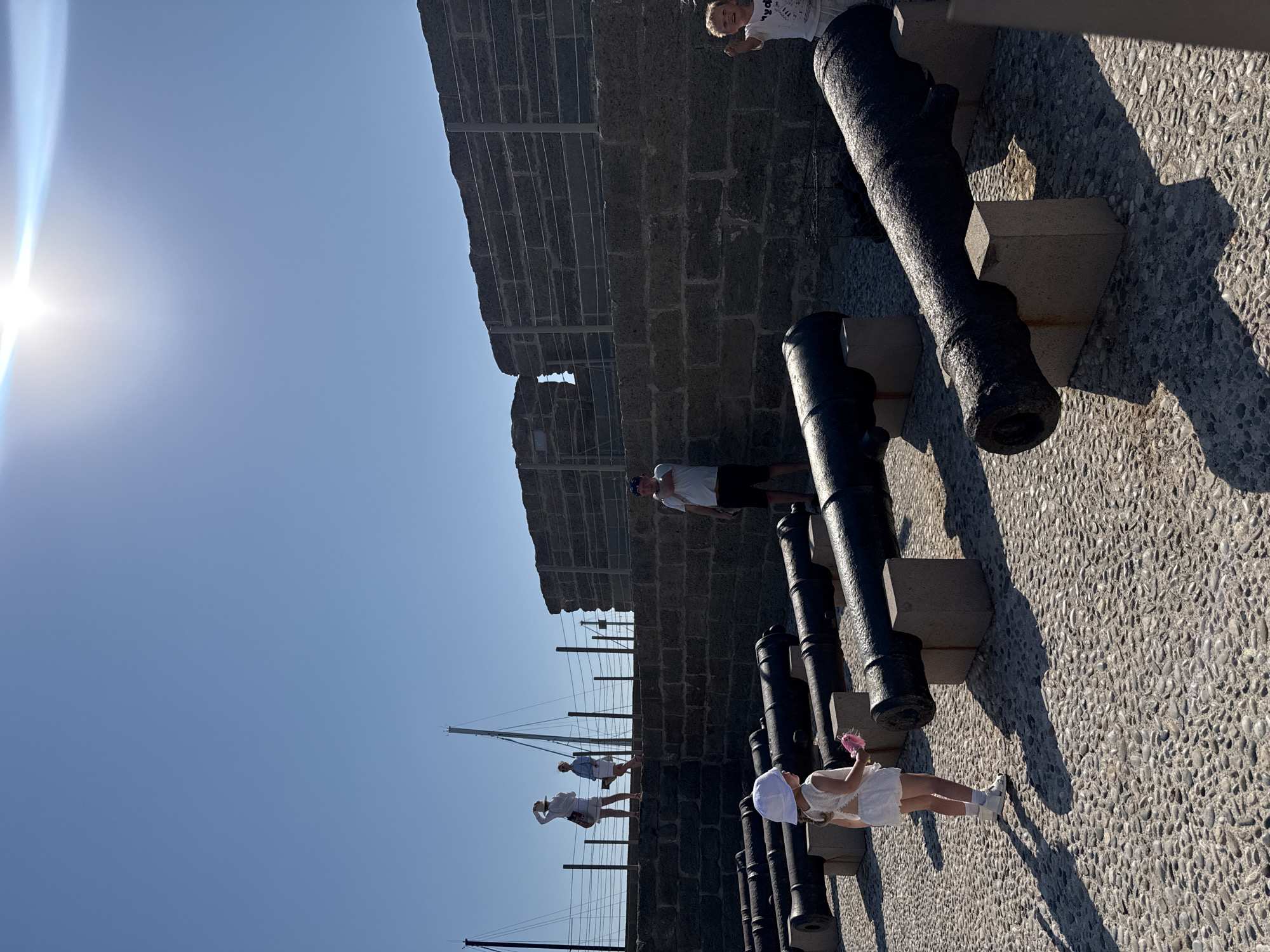 In Bodrum, Turkey, on a sunny day, a row of antique cannons are displayed on a cobblestone ground in front of a stone wall. A young girl in a white dress and hat stands near the cannons, holding a pink toy. Further back, a man in a white t-shirt and shorts observes the scene. Two women and another child are visible on top of the stone wall, looking out towards the sea. Boats and masts can be seen in the distance. The sun is bright in the clear blue sky.