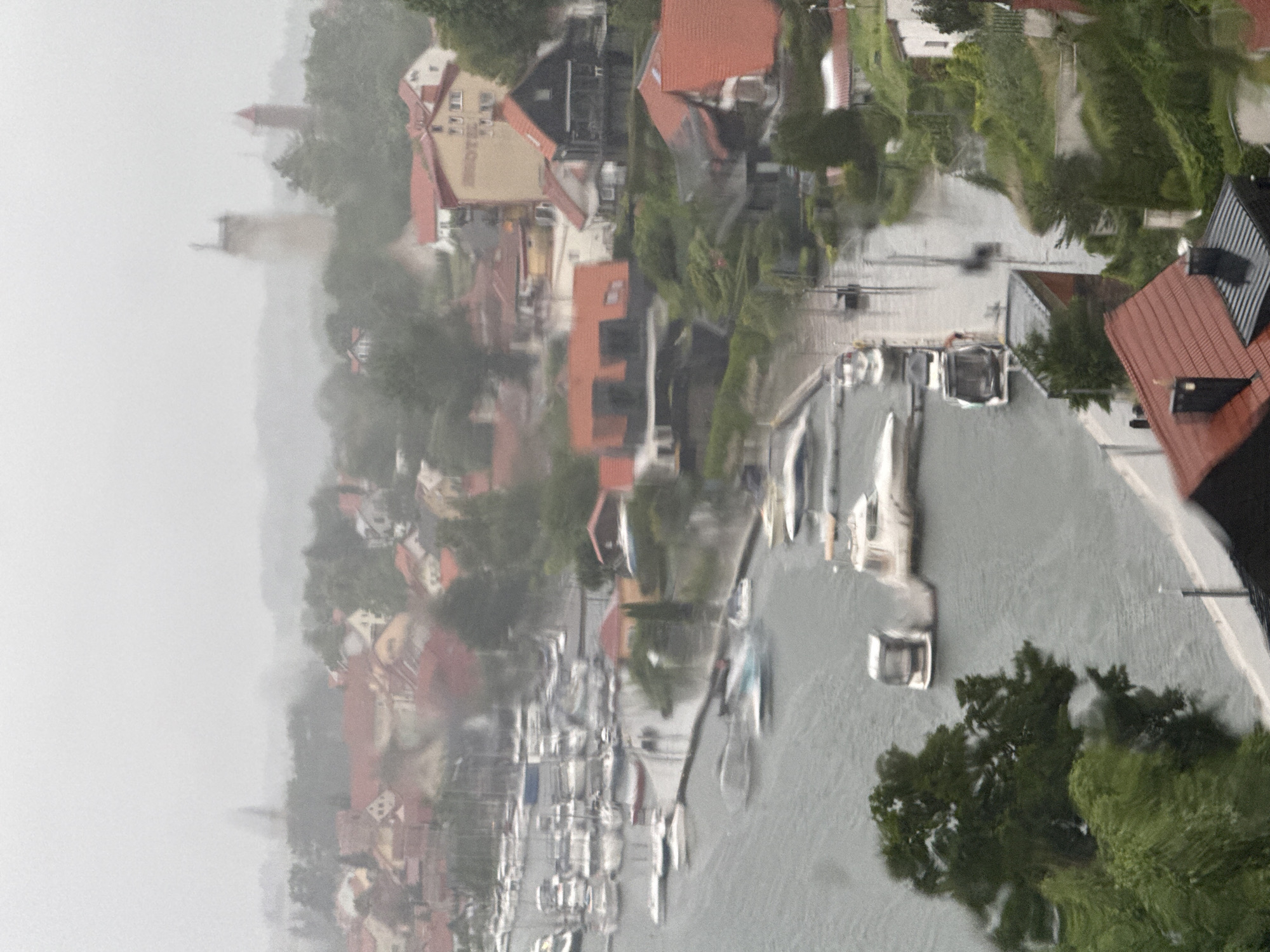 The image depicts a hazy, overcast day in the city of Mikołajki, Poland, as indicated by the faint outline of a tower visible in the distance. The foreground is dominated by a marina filled with numerous boats of varying sizes, docked and seemingly at rest. The water surface appears choppy, reflecting the moody atmosphere. Beyond the marina, the cityscape unfolds with a cluster of buildings characterized by red-tiled roofs. Some of these structures appear to be residential homes, while others, like the taller building with multiple windows and the word "HOTEL" visible, suggest commercial establishments. Lush green trees are interspersed throughout the urban landscape, adding a touch of nature to the scene. The overall impression is one of a tranquil, perhaps even slightly somber, day in a waterfront town.