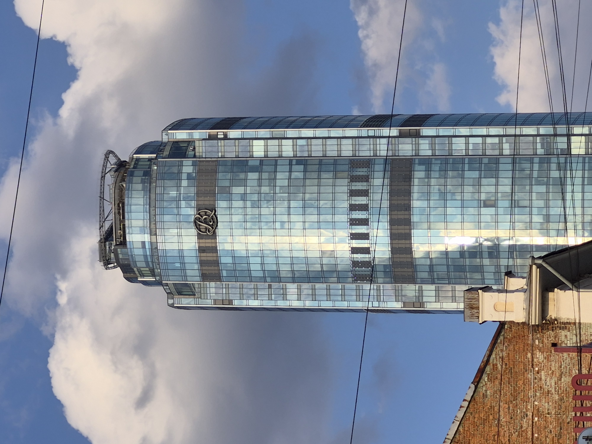 The image shows the Vysotsky business center, a skyscraper located in Yekaterinburg, Russia. The building, with its distinctive blue glass facade, is the central subject of the photograph. It features a helipad at its summit and a prominent "B" logo on its upper section. The skyscraper is partially obscured by a brick building in the foreground, which has a red awning and some architectural details. Power lines traverse the scene horizontally, creating a visual interruption. The sky above is partly cloudy, with patches of blue sky visible amidst large, white clouds. The lighting suggests it is daytime, with sunlight reflecting off the glass surfaces of the skyscraper.