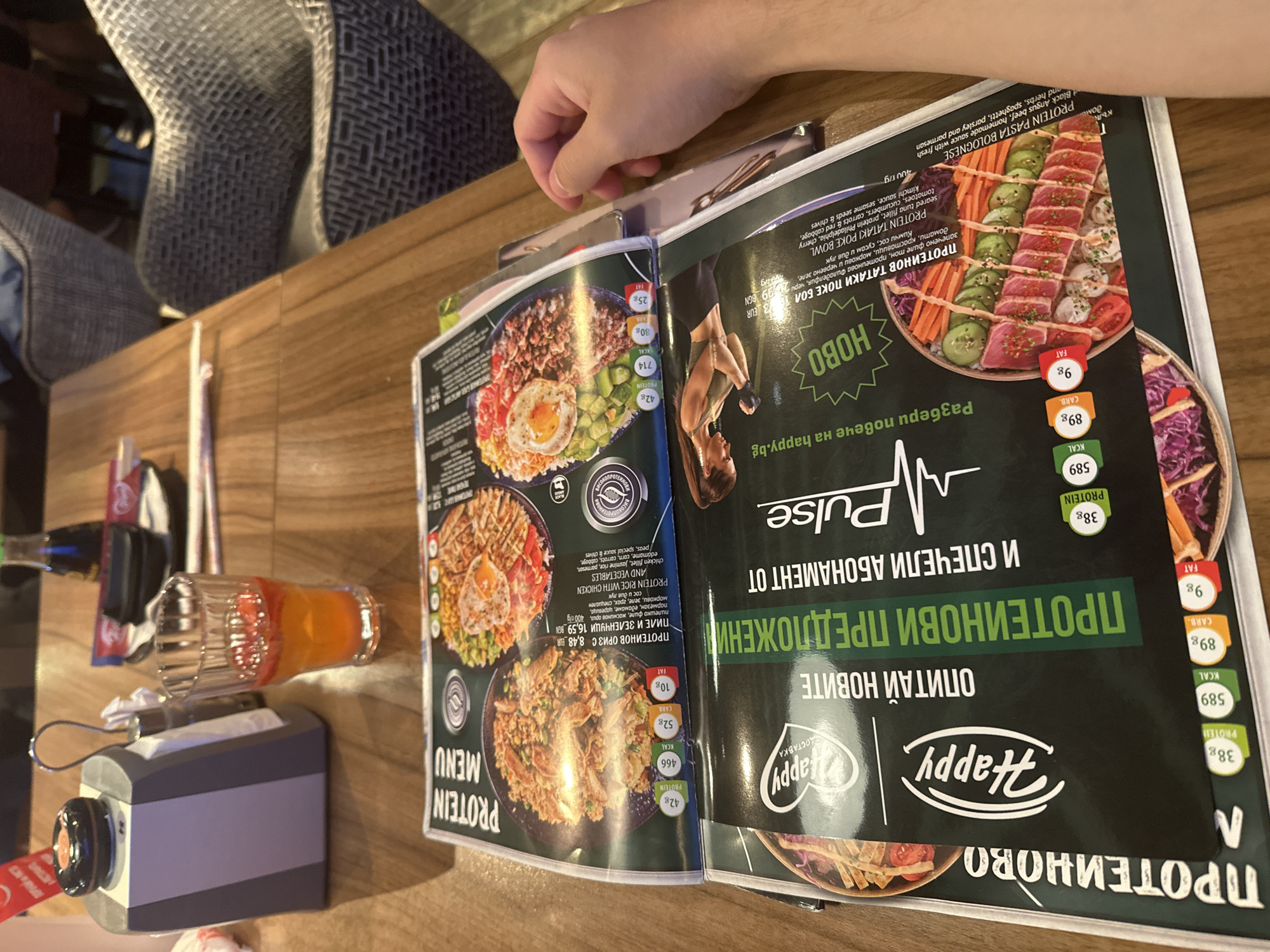 A close-up, high-angle shot captures a restaurant table in Unknown, Unknown, with a person's right hand visible on the right side. The table is made of light brown wood with visible grain.
Two open menus are prominently displayed, stacked one on top of the other, lying flat on the table. The top menu, labeled "PROTEIN MENU" in English, features colorful photographs of various protein-rich dishes. Visible details on this menu include "PROTEIN RICE WITH CHICKEN AND VEGETABLES" with nutritional information showing "42g PROTEIN," "466 KCAL," "52g CARB," and "10g FAT" for one dish, and "42g PROTEIN," "714 KCAL," "30g CARB," and "25g FAT" for another. Bulgarian text is also present on the menu, describing ingredients and prices (e.g., "ПРОТЕИНОВ ОРИЗ С – 8.48 EUR" and "ПИЛЕ И ЗЕЛЕНЧУЦИ 16.59 BGN"). The bottom menu, partially visible, displays a large "Happy" logo and also features protein-focused dishes, with text in Bulgarian including "ОПИТАЙ НОВИТЕ ПРОТЕИНОВИ ПРЕДЛОЖЕНИЯ" (Try the new protein offers) and "И СПЕЧЕЛИ АБОНАМЕНТ ОТ Pulse" (and win a subscription from Pulse). A photo of a woman in athletic wear is on this menu, alongside a "Pulse" logo.
To the left of the menus, a grey and white napkin dispenser is present, with a black circular object, possibly a call button or pager, on its top. A small glass of an orange-colored drink with red fruit pieces (possibly strawberries) floating on top is positioned to the right of the napkin dispenser. In the background, slightly out of focus, a dark glass bottle, likely soy sauce, and a small white plate with chopsticks rest on the table. Beyond the table, two woven, patterned grey chairs are visible, suggesting a dining environment. The overall lighting is warm and even.