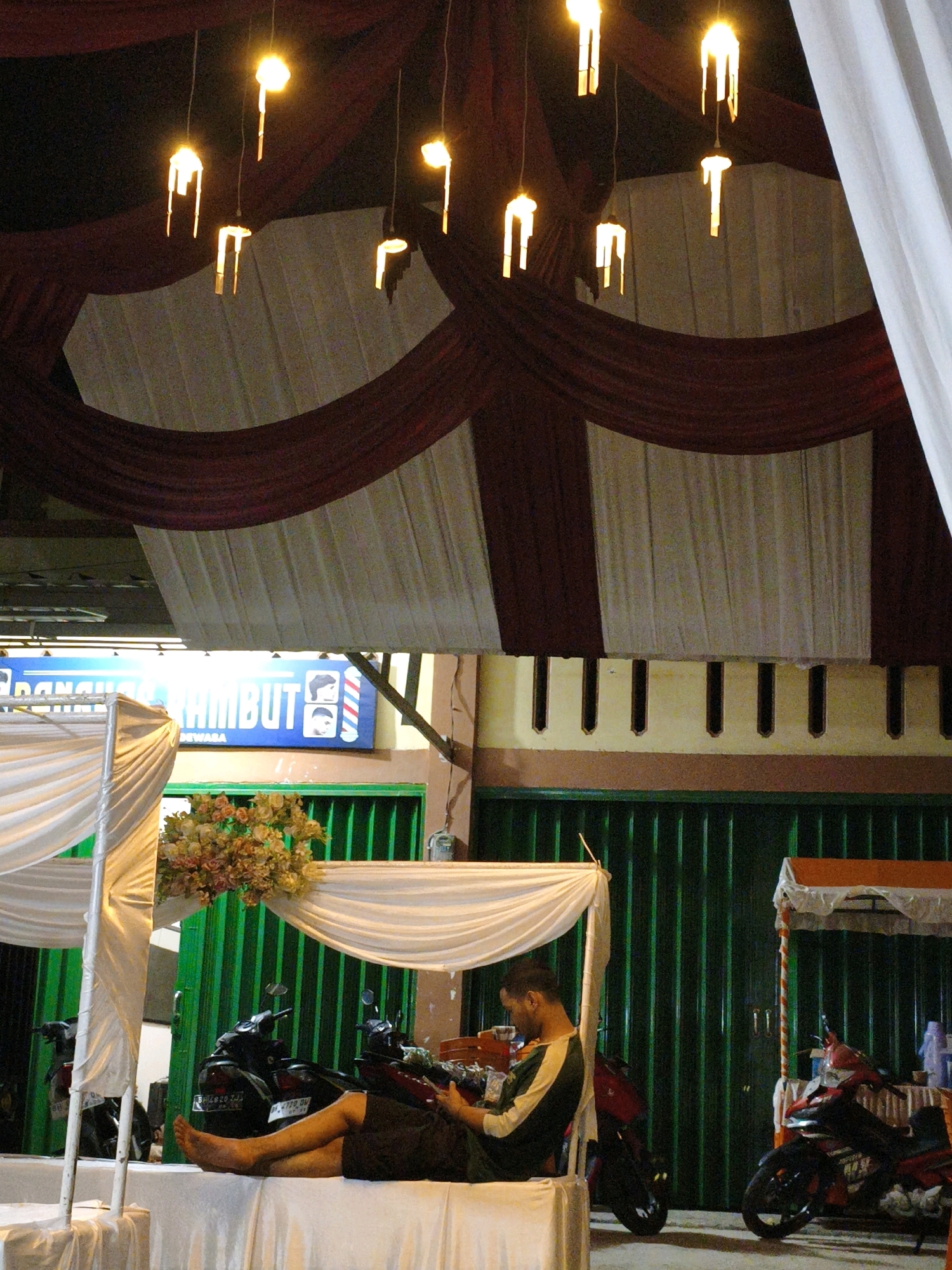 The image depicts an outdoor setting at night, possibly a street or an event space, in Koto Baru Semurup, Indonesia. The scene is illuminated by decorative lights, with several unique, candle-shaped fixtures hanging from above. These fixtures are fashioned from cylindrical elements with openings, emitting a warm, yellow light.
Dominating the upper portion of the image is a draped canopy made of dark red and white fabric, creating a festive or ceremonial atmosphere. Below this, a blue sign with white lettering reads "BARBER RAMBUT" (Barber Hair), featuring stylized depictions of a man's haircut and a barber pole, indicating the presence of a barbershop or a related business.
In the middle ground, there is a person, appearing to be a man, seated with his legs extended. He is wearing dark shorts and a shirt with green and yellow stripes. He is looking down, possibly at a device in his hands. In front of him, a white draped table serves as a surface. Behind him, several motorcycles are parked. One is a red and black scooter with license plate "BH 2828 ZJJ." To the right of this, another motorcycle is visible.
The background features a corrugated green metal gate or door. A small stall with a white canopy and orange trim is positioned to the right, with another motorcycle parked in front of it. A bouquet of flowers in shades of pink, yellow, and green is displayed above the green gate. The overall impression is of a lively, community-oriented area, possibly during an evening gathering or market.