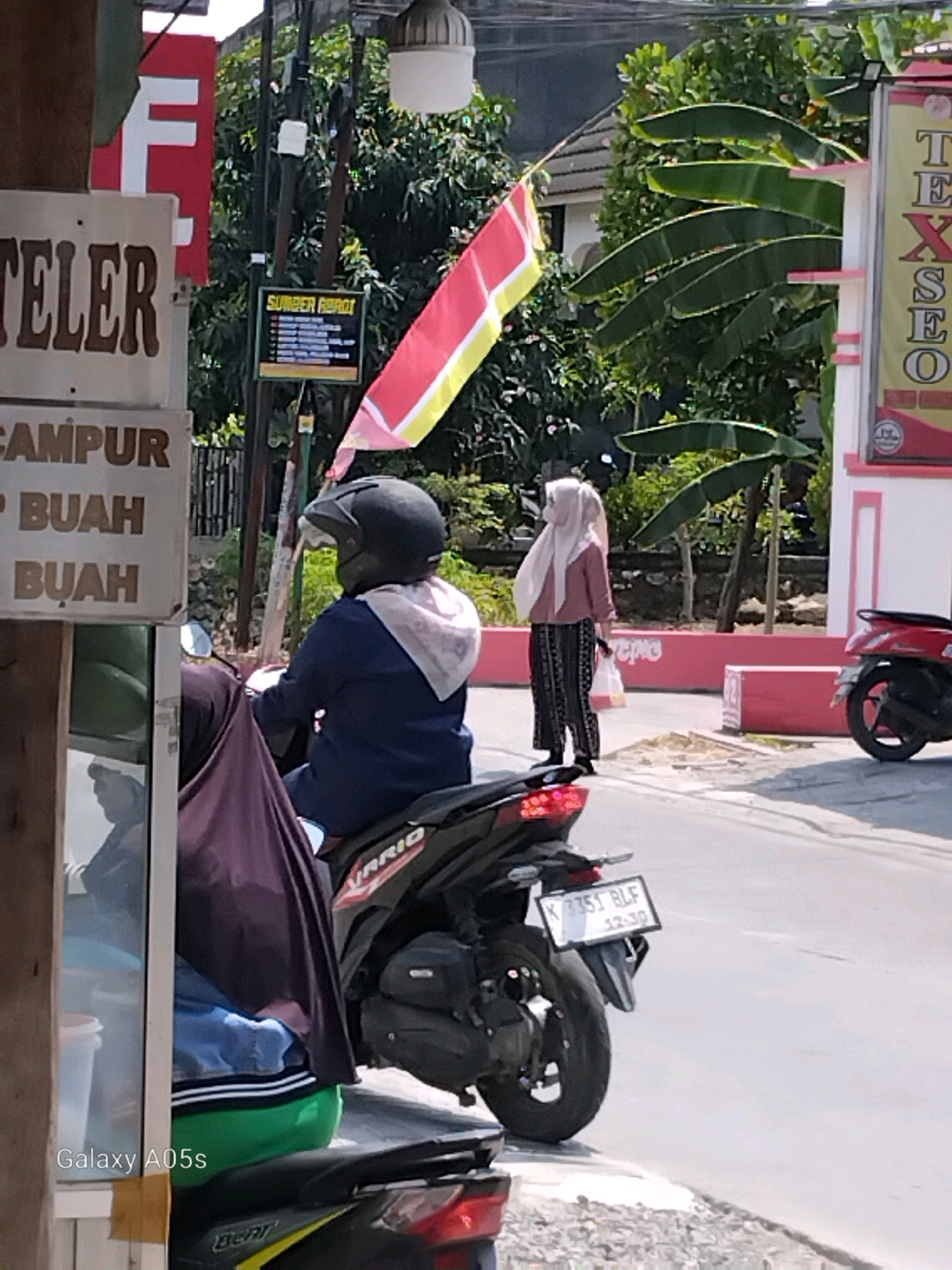 The image depicts a street scene in Karangayung, Indonesia, under bright daylight. In the foreground, a person wearing a dark blue hoodie and a light-colored hijab is seated on a motorcycle, facing away from the camera. Another person, also wearing a hijab, is standing to the right, holding a white plastic bag. A red motorcycle is parked on the right side of the road.
Various signs are visible in the background. One sign on the left, with a red background and white lettering, displays the letter "F". Below this, a white sign has brown text that reads "TELER" followed by "CAMPUR BUAH BUAH". A smaller yellow sign with black text, labeled "SUMBER ABADI", is also present. On the far right, a tall, vertical sign has the word "TEXSEO" written in black letters.
The environment includes trees with large leaves, a building with a light-colored facade and pink trim, and a paved road. The overall atmosphere suggests a commercial or residential area in a town or city.