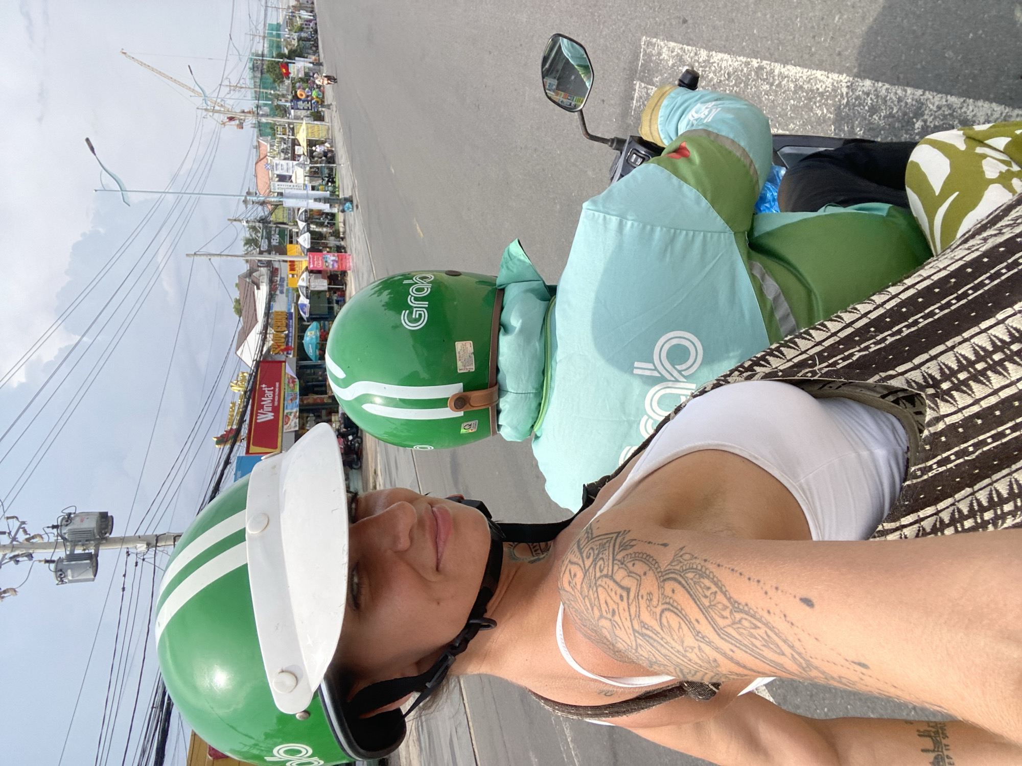 A woman with a green and white helmet and a white tank top is riding on the back of a motorcycle in Phu Thuy Ward, Vietnam. She has a large tattoo on her left arm and is carrying a patterned bag. The motorcycle driver, wearing a light blue and green jacket with the "Grab" logo, is driving the motorcycle down a street lined with shops and power lines. The sky is cloudy.