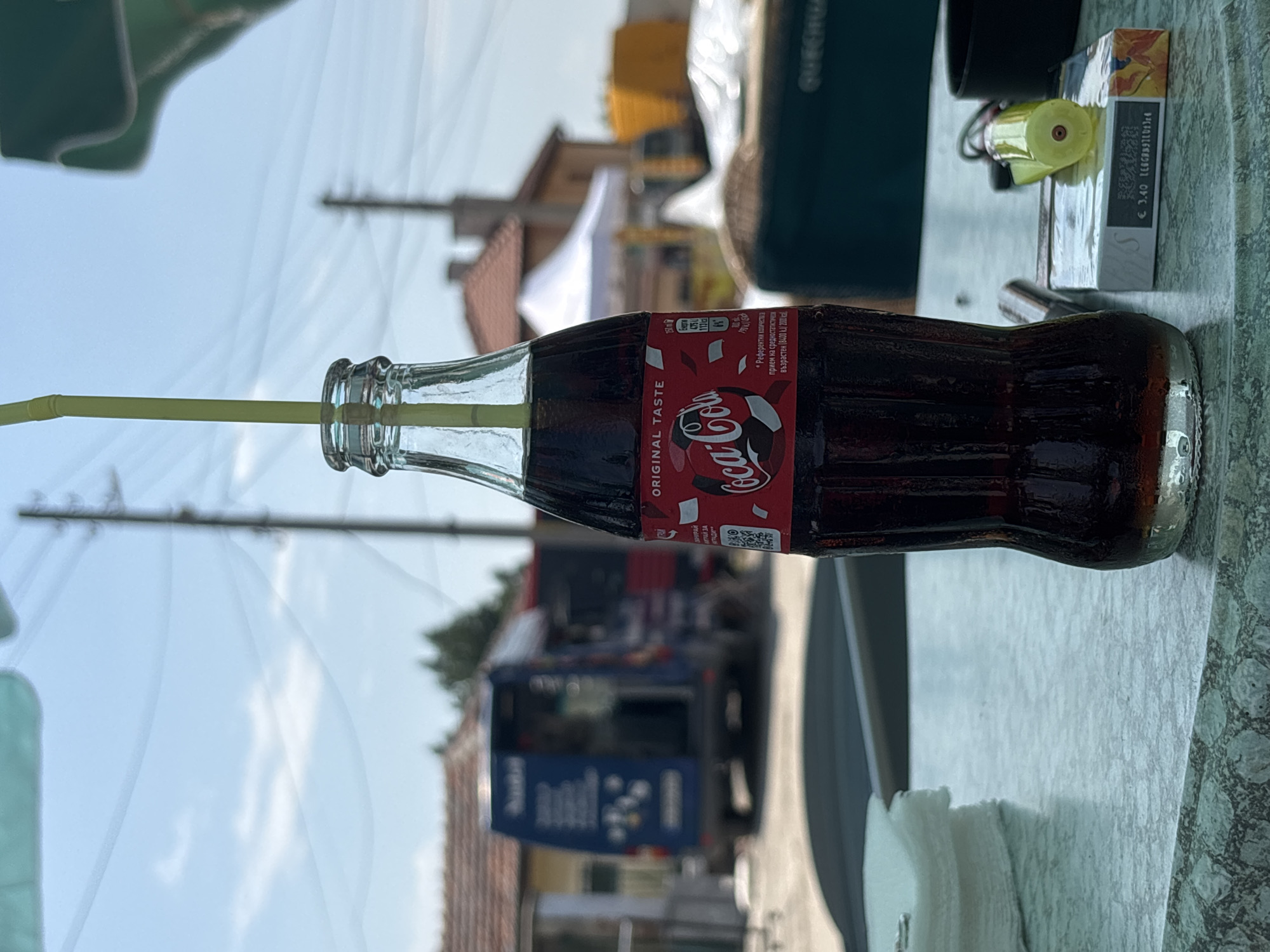 A clear glass bottle of Coca-Cola, nearly full with dark brown liquid, stands centrally on a light green mottled table, featuring a yellow plastic straw inserted into its opening. The bottle's red label displays "ORIGINAL TASTE" and the "Coca-Cola" logo in white script, along with a graphic of a soccer ball and nutritional information in Cyrillic, indicating "250 ml" and "475 kJ / 113 kcal." To the left of the bottle, a stack of white paper napkins is visible. To the right, a white pack of "L&S" branded cigarettes, marked with a price of "€ 3.40" and additional Cyrillic text, rests on the table next to a yellow disposable lighter and a partially visible black ashtray.
The background is an outdoor, sunlit environment, blurred, under a clear blue sky with sparse white clouds. Utility poles and overhead wires are visible against the sky. Distinct elements in the midground include a dark blue utility or service van, positioned to the left of the bottle's center, with its rear doors open, revealing internal shelving or equipment. White text or graphics are present on the van's side, possibly including the word "BARO." Beyond the van, brown tiled roofs of buildings are visible. To the right of the blue van, a white canopy tent structure is present, with a yellow or orange vehicle partially visible further in the distance. The scene is located in Kostievo, Bulgaria.