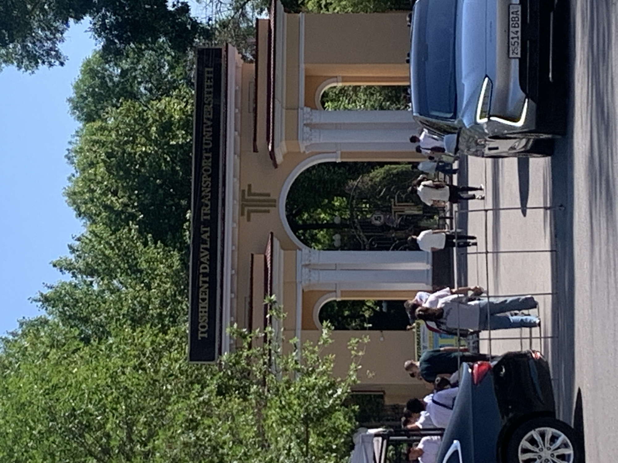 The image shows the entrance to the Tashkent State Transport University in Tashkent, Uzbekistan. A sign with the university's name is visible on the left side of the entrance arch. Several people are walking towards the entrance, and cars are parked on the side of the road. The environment includes trees and sunlight.