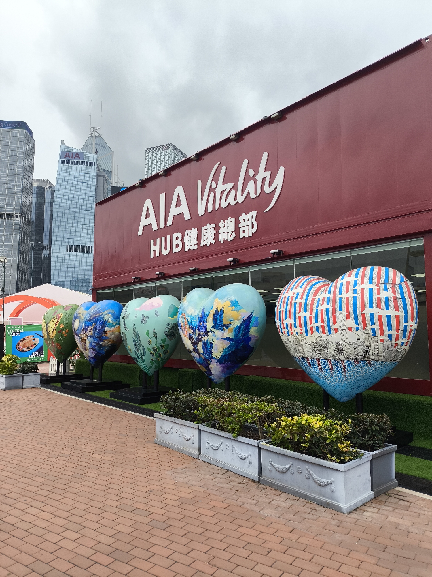 The image displays an outdoor public art installation and event space in the Central Harbourfront area of Hong Kong, China, on an overcast day. Five large, heart-shaped sculptures are arranged in a linear fashion in the mid-ground. From right to left, the sculptures feature: a red and blue striped pattern over a cityscape silhouette on a white background with a blue mosaic base; a teal background with green foliage and light pink butterflies; a dark blue background with an intricate painting of a large bird with outstretched wings; a lighter blue background with a moon and an abstract, whimsical scene; and a green background with a large yellow butterfly and floral elements. Each heart sculpture rests on a dark rectangular base, situated on artificial green turf.
Behind the sculptures stands a maroon-red, rectangular building prominently displaying "AIA Vitality HUB 健康總部" in white lettering. The building's lower section features horizontal window panes.
In the foreground, a reddish-brown brick paved walkway extends from the bottom of the frame. Along the right side of this walkway, several grey rectangular planters adorned with garland-like motifs contain green shrubbery. To the far left, a portion of an orange and white tent structure is visible, alongside a green banner advertising "CENTRAL YARDS" and an "EDIBLE ART FAIR" with dates "2024.03.30-04.05".
In the background, under a grey, cloudy sky, several prominent Hong Kong skyscrapers are visible, including the AIA Central building, the Bank of China Tower, and other high-rise structures, confirming the urban setting in Central, Hong Kong Island. No individuals are present in the scene.