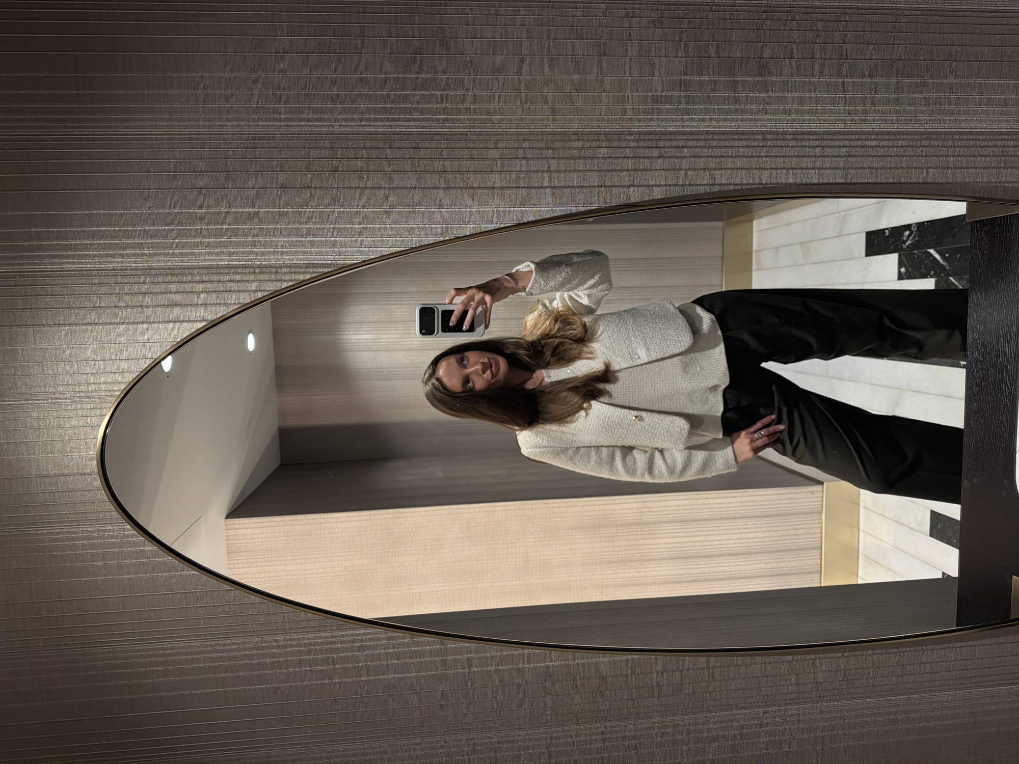 A woman is captured taking a mirror selfie in an interior space, identified as being in Budva, Montenegro. The central subject, a woman with long brown hair, is dressed in a white, textured, cropped jacket featuring gold-colored buttons, worn over a lighter-colored top, and dark, wide-leg trousers. A silver-toned ring is visible on her right hand, and a light-colored bracelet adorns her left wrist. She holds a white foldable smartphone, consistent with a Samsung Galaxy Z Flip model, with her left hand. Her head is slightly tilted, and her gaze is directed towards the upper left, away from the phone's camera lens.
The mirror is an arch-shaped, full-length fixture, framed by a thin gold-colored border, with its lower section resting on a dark-toned base. The wall visible outside the mirror on the right side of the image is covered in a dark, vertically textured wallpaper. Within the mirror's reflection, the interior walls are a lighter beige hue, also featuring a vertical textured pattern. The reflected ceiling is white and includes two visible recessed downlights. The floor, reflected in the lower portion of the mirror, is composed of white and black tiles arranged in a geometric pattern, indicative of a polished surface like marble. The overall environment suggests an upscale indoor setting.