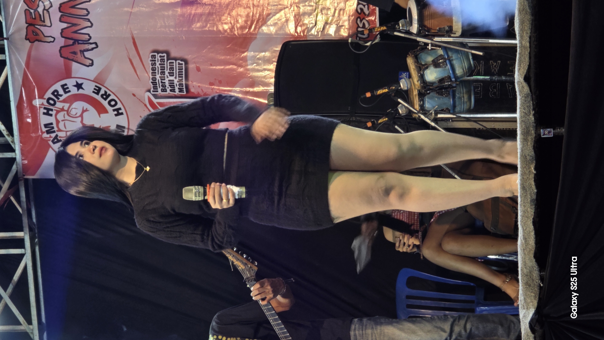 A woman stands on a stage, holding a microphone and wearing a dark, fitted dress and a dark sweater. She has dark hair and is looking to her right. A banner with red and white elements and text is visible behind her. To her left, a person is playing a guitar. In the background, there are speakers and drums. The stage appears to be set up for a performance. The image also includes a watermark in the lower left corner that reads "Galaxy S25 Ultra". The text "Juwana, Indonesia" is part of the background banner.