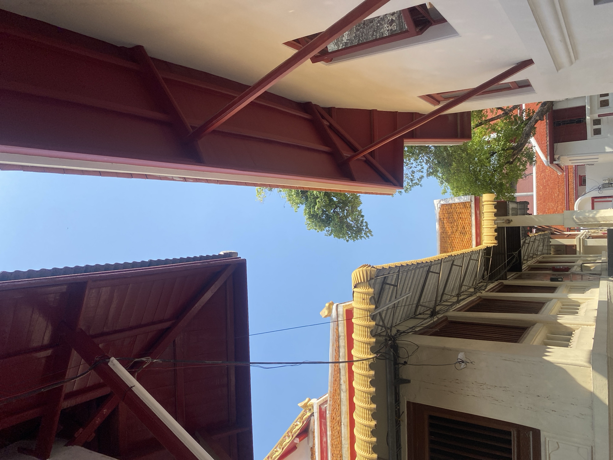 A low-angle shot in Bangkok, Thailand, shows a view of rooftops and the sky. Red-painted wooden roofs are visible on the left and top of the frame, with support beams. The building on the right is taller, featuring a corrugated metal roof and yellow decorative elements. A few trees are visible in the gaps between the buildings. The sky is clear and blue. Electrical wires run across the view between the buildings.