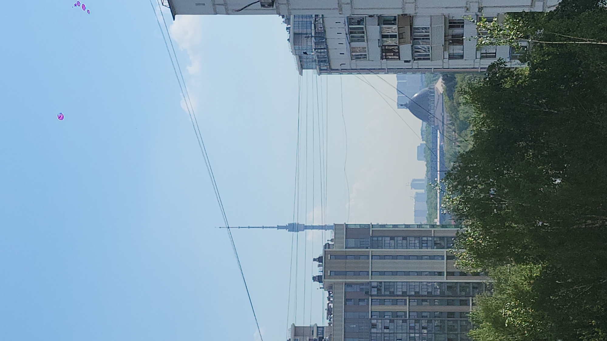 The image shows a cityscape with a prominent television tower, likely the Ostankino Tower in Moscow, Russia. In the foreground, there are residential buildings, some appearing modern with multiple windows and others appearing older with balconies. The sky is light blue with a few scattered balloons in the upper left. High-voltage power lines stretch across the view. There are trees in the background. The cityscape appears hazy, indicating possible air pollution or fog.