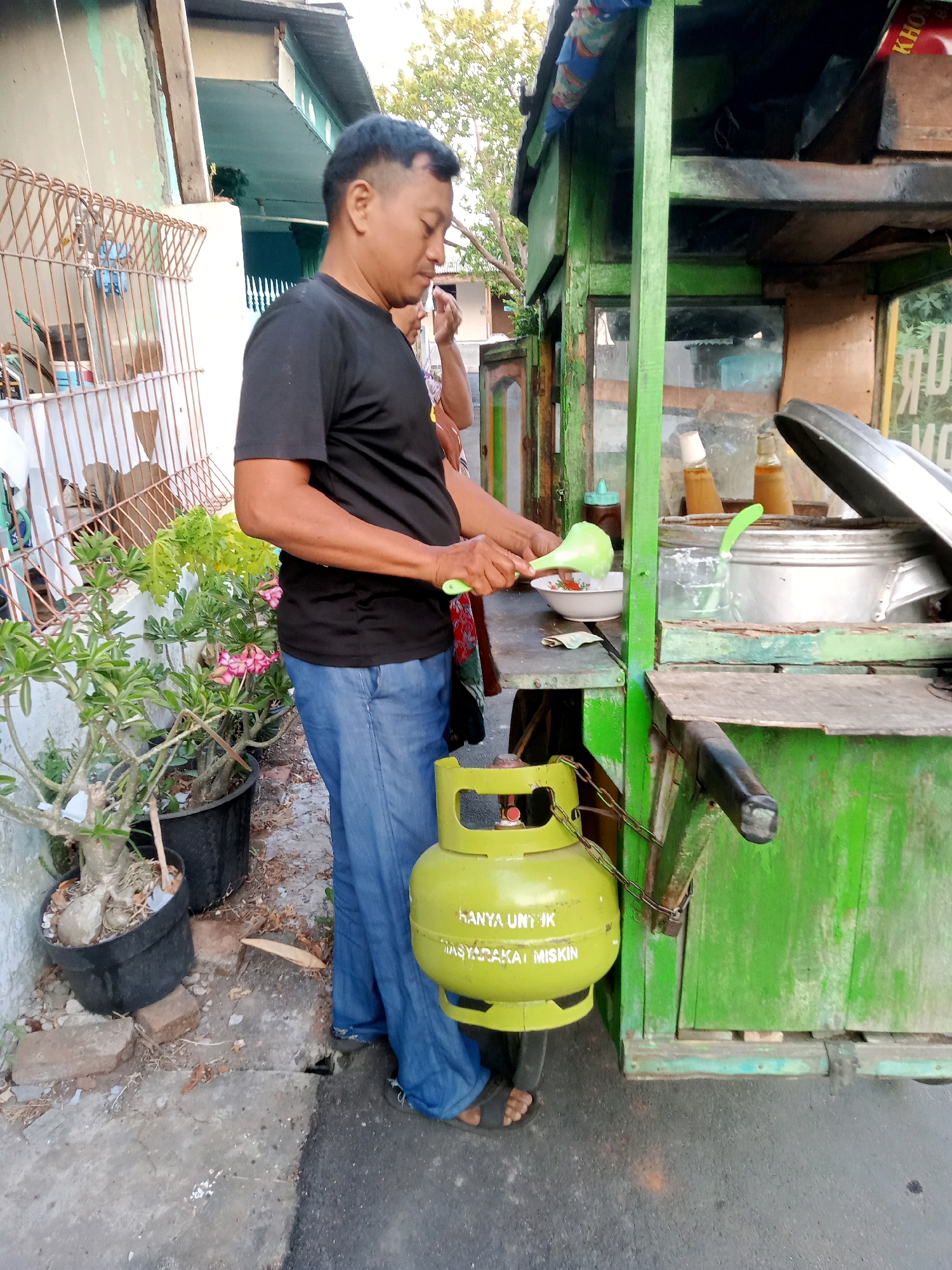 A man in a black t-shirt and blue jeans stands next to a green food cart, holding a green ladle and serving a bowl. A yellow LPG gas cylinder is attached to the cart by chains. The cart appears to be a street food vendor, common in Cirebon, Indonesia. The background shows a residential building with a tiled roof and some trees. The area seems to be an outdoor setting, possibly a sidewalk or street.