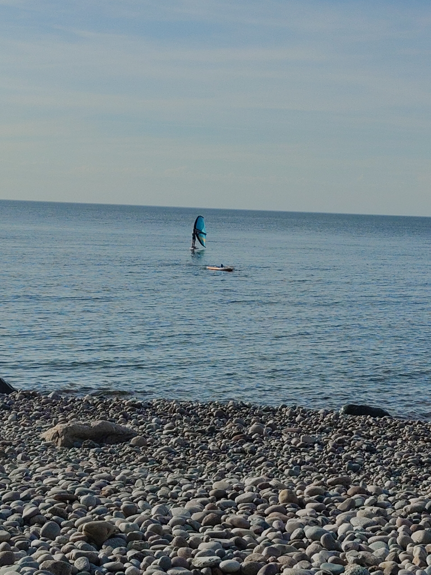 In Pionersky, Russia, a person is wing foiling on the calm, blue Baltic Sea. They are standing on a board, holding a bright blue wing. The water is slightly rippled, and the sky above is a pale blue with a hint of haze. In the foreground, a pebble beach stretches along the shoreline, covered in smooth, grey and tan stones of various sizes. The scene is tranquil, with the person and their water sport activity being the sole focus in the expansive seascape.