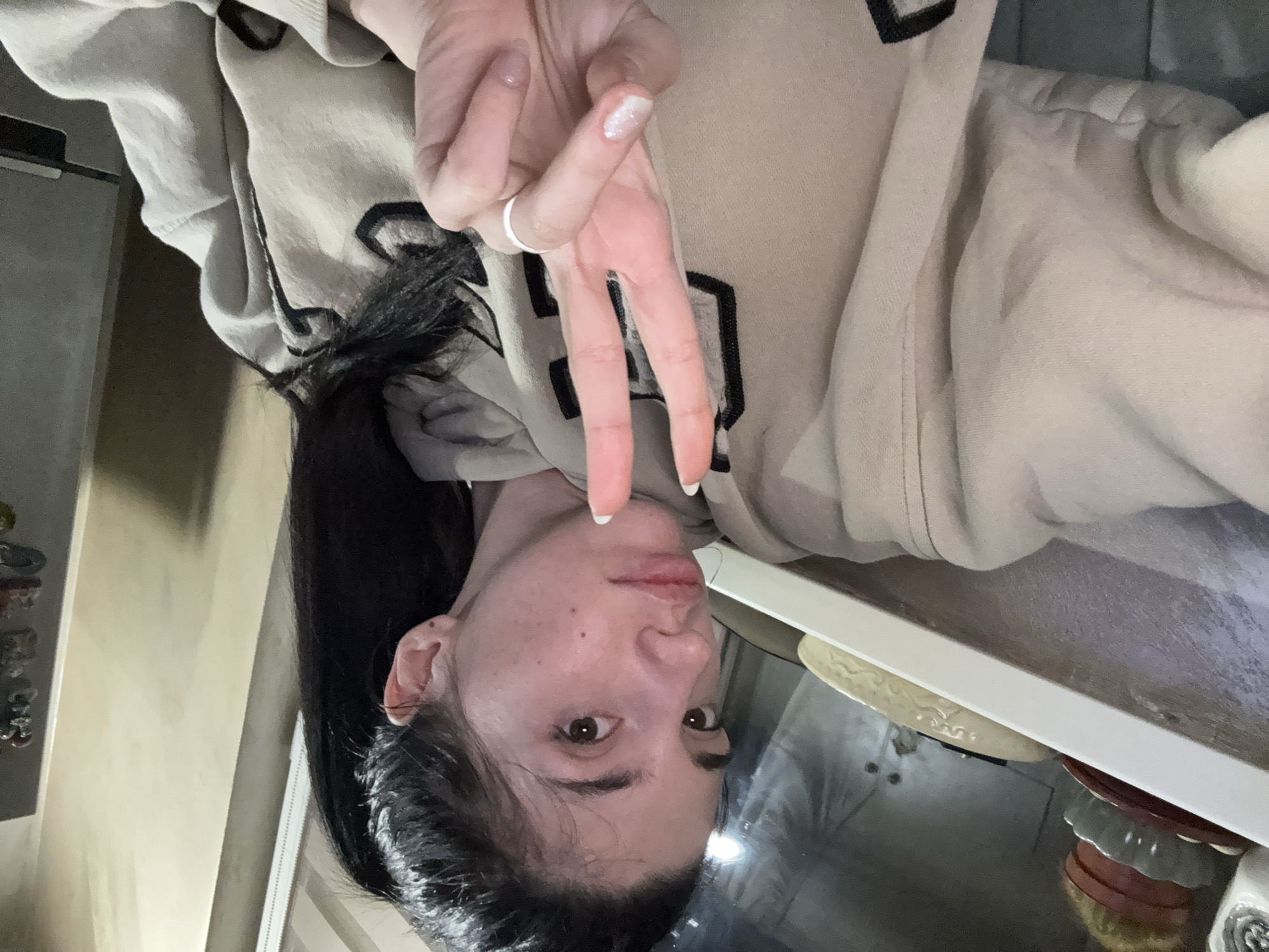 A young adult woman with long dark hair is positioned in the lower left quadrant of the image. She is wearing a light beige hoodie with dark block letters on the front. Her face is visible, displaying light skin with blemishes, dark eyebrows, and brown eyes. She is making a peace sign with her right hand, which is in the upper center of the image. She is wearing a ring on her ring finger. The background shows a countertop with a stack of decorative plates in the lower right corner. To the left, part of a wall is visible with decorative items hanging. The image is taken indoors, presumably in an apartment or house. The location within the city of Sofia, Bulgaria, cannot be determined from the visual information provided.