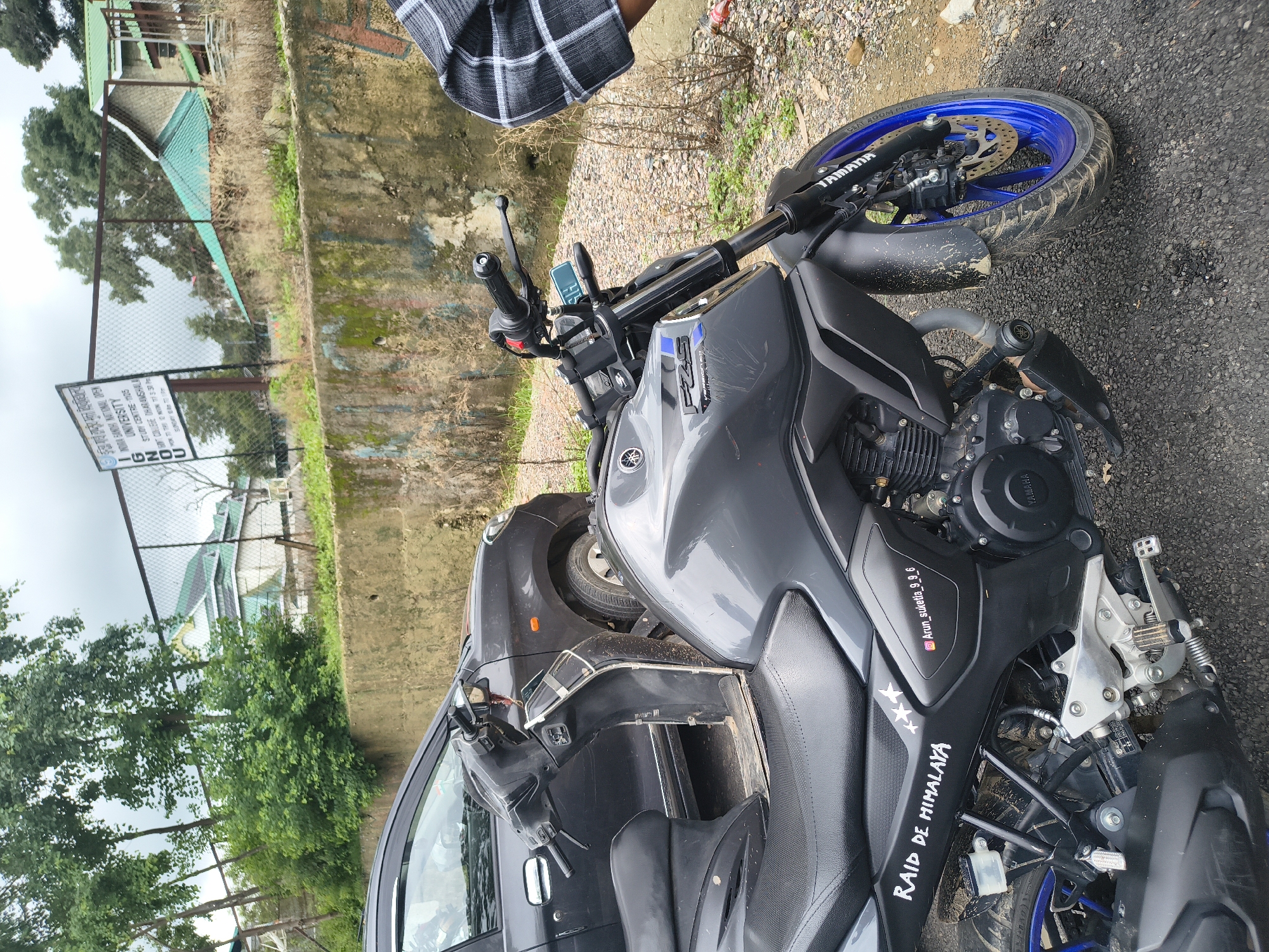 An exterior, medium shot captures a dark gray motorcycle parked next to a dark gray car. The motorcycle is in the foreground, angled towards the viewer, with its front wheel and part of its engine visible. The car is parked behind the motorcycle, on its left side. The scene is set on a road with gravel and some green vegetation. In the background, a wire fence separates the road from a concrete wall and some buildings with green roofs. A signpost is visible near the fence. The sky is overcast.