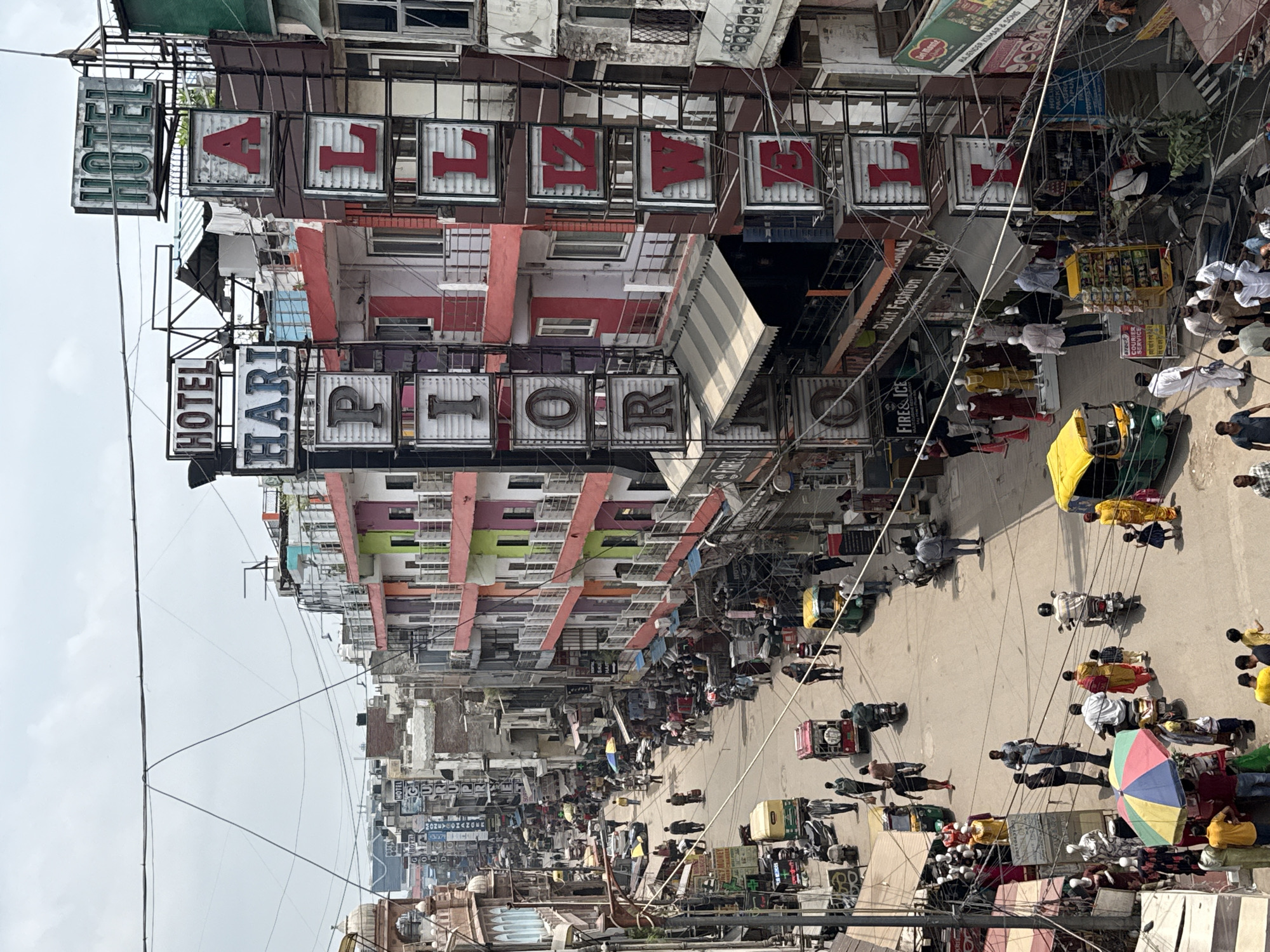This is an elevated, medium-angle, eye-level shot of a bustling street scene in Delhi, India, likely from a higher vantage point such as a building or overpass. The street is lined with multi-story buildings, many of which feature colorful facades and prominent hotel signage. The architecture appears to be a mix of older and more modern styles.
The street itself is paved and appears to be busy with a variety of modes of transportation, including auto-rickshaws, scooters, and motorcycles. Pedestrians are also abundant, walking along the sidewalks and crossing the street. Many people are dressed in casual clothing, with some men wearing shirts and pants, and women in what appears to be traditional Indian attire like salwar kameez.
Numerous shops and businesses are visible along the street, with awnings extended and merchandise displayed. Signs for "HOTEL HARI" and "HOTEL ALIZW" are prominently displayed on the buildings, indicating lodging establishments. There are also signs for businesses like "Dixit Fashion," "Fire & Ice," and various other shops with Indian script. The overall impression is one of a vibrant, commercial district.
The sky is overcast, suggesting a cloudy day. There is a complex network of electrical wires and cables crisscrossing the scene, a common feature in urban Indian environments. The scene is dynamic, with people and vehicles in motion, creating a sense of ongoing activity.