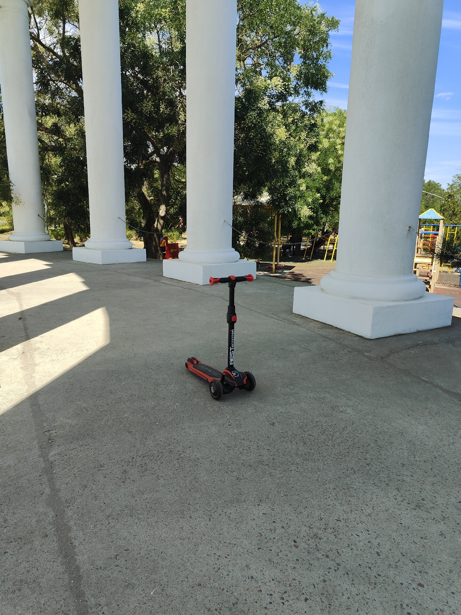 A three-wheeled scooter with red handlebars and red accents on the wheels sits on a paved surface, possibly in Chornomorsk, Ukraine, near a row of large white pillars. Green trees and a blue sky are visible in the background, along with what appears to be a playground structure. The scooter is the primary subject in the foreground.