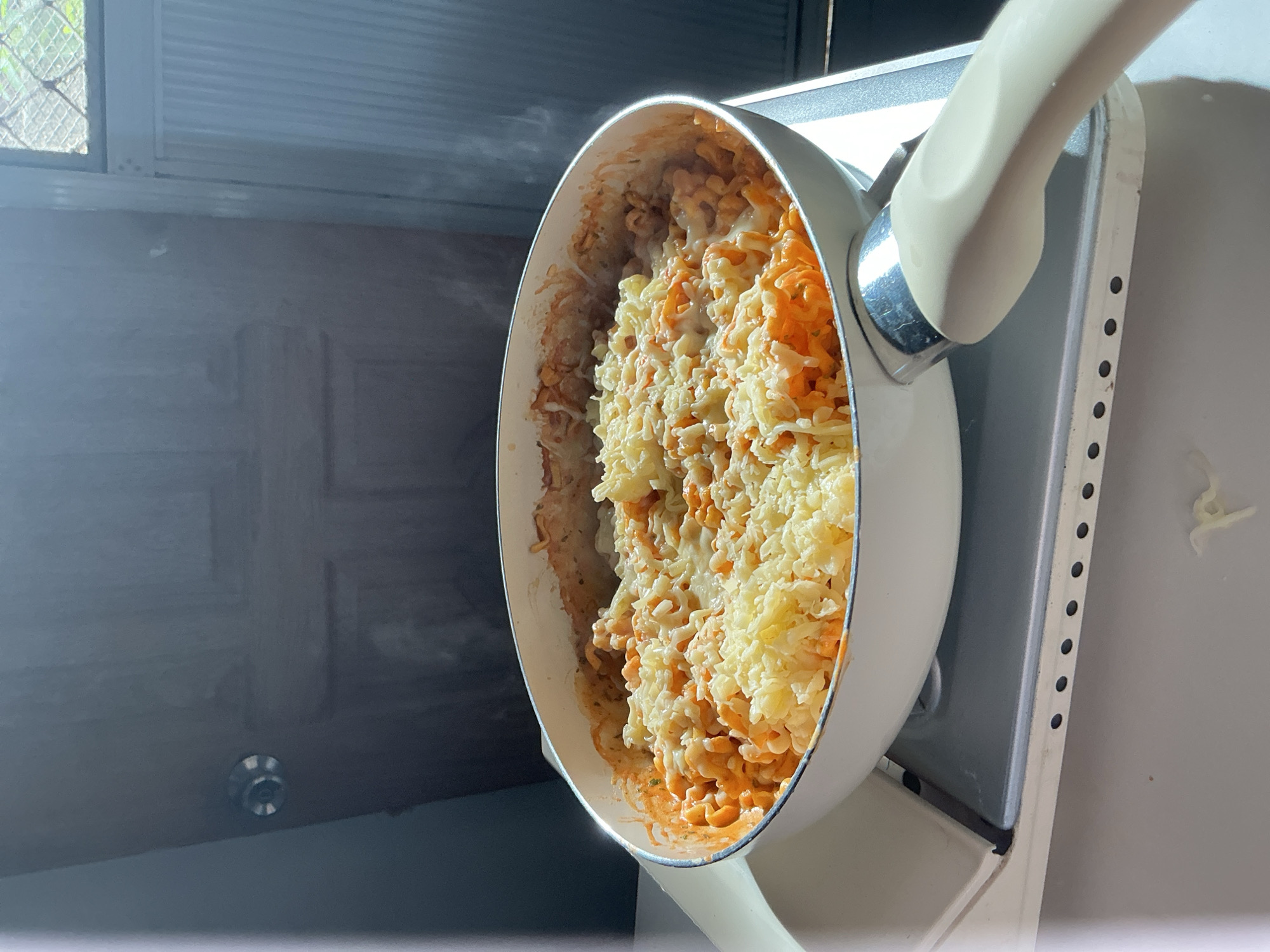 The image shows a white-handled pan filled with pasta covered in melted cheese and tomato sauce. The pan is placed on a stovetop. Steam is rising from the pasta. The background appears to be a room with a closed door and a window with a diamond pattern. The scene suggests a domestic setting where food is being prepared or served. The reference to Davao City, Philippines, is not visually supported by the image content.