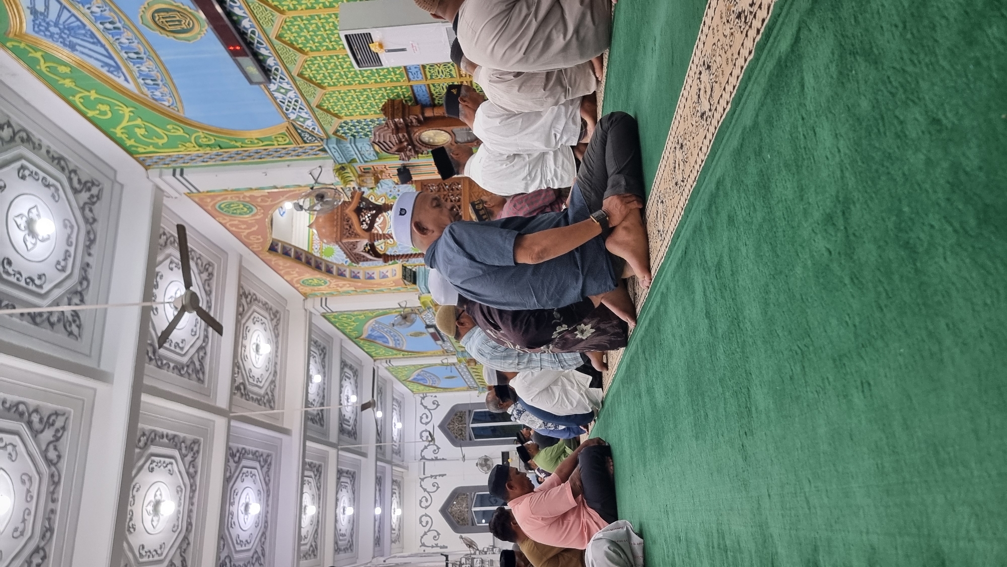 The image depicts a group of men engaged in prayer inside a mosque. They are seated on a green carpet with a patterned border, facing a central point of focus, likely the mihrab or qibla wall. The men are dressed in traditional attire and prayer caps. The mosque's interior features ornate ceilings with intricate patterns and lighting fixtures, as well as colorful wall decorations with geometric designs and calligraphy. In the background, a wooden pulpit and a large clock are visible. The overall atmosphere suggests a religious gathering. The location is identified as Keudekruenggeukueh, Indonesia.
