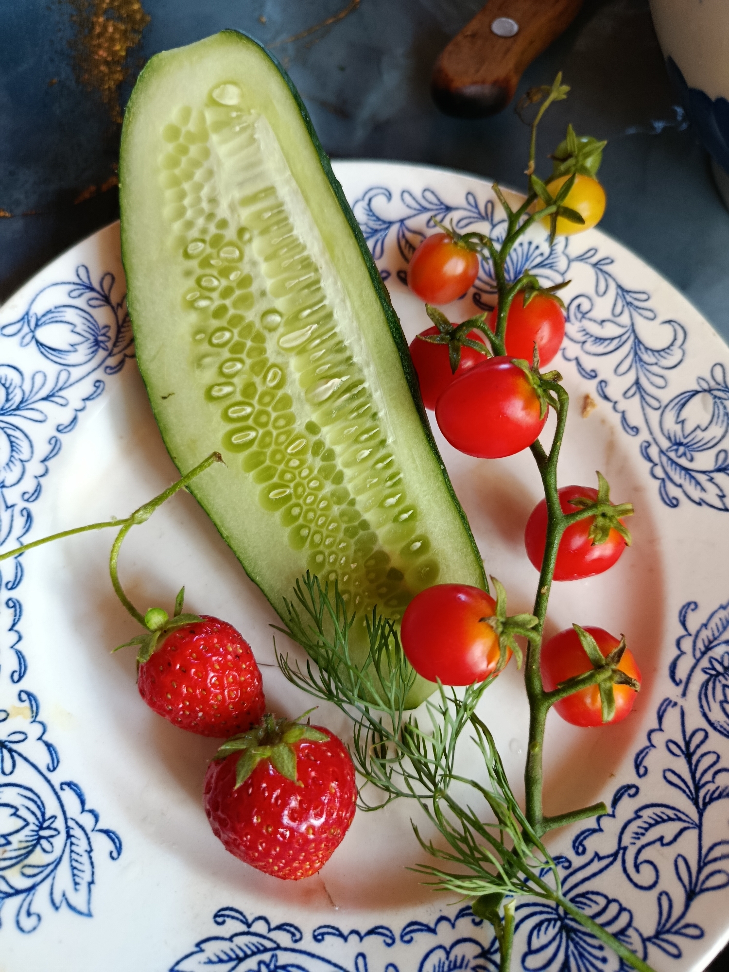 The image displays a close-up of a white plate adorned with a blue floral pattern. On the plate are several food items: a halved cucumber, a cluster of cherry tomatoes on the vine, two strawberries, and a sprig of dill. The cucumber is sliced lengthwise, revealing its seedy interior. The cherry tomatoes are a mix of red and yellow, with some appearing slightly unripe. The strawberries are bright red and appear fresh. The dill is a delicate green herb. The background is dark and textured, with hints of gold. The scene is lit from above, casting soft shadows. There is no indication of the city of Верея, Russia, or any specific location within it in the provided image.