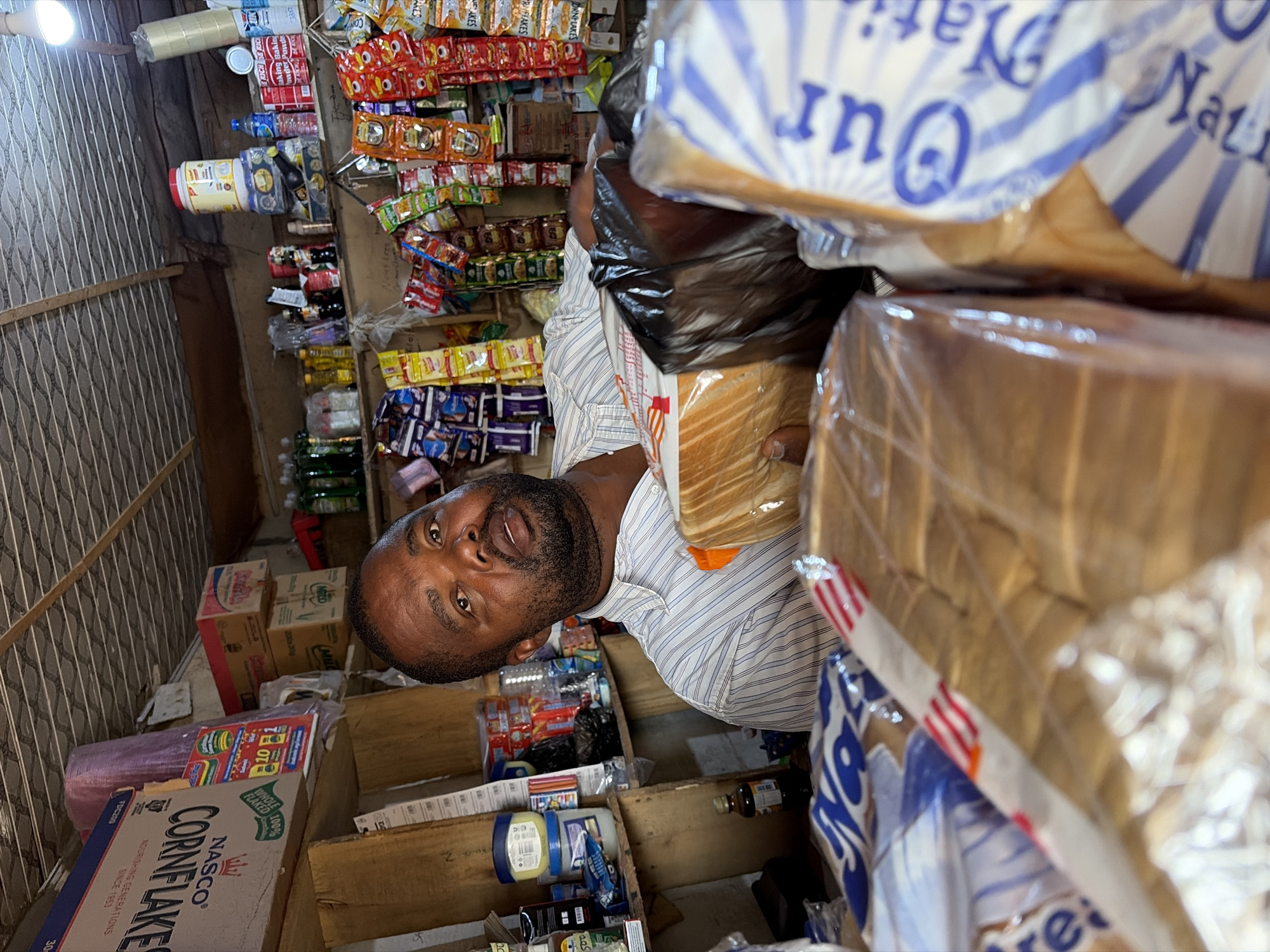 In what appears to be a small shop in Jos, Nigeria, a bearded man is seen holding a loaf of bread wrapped in plastic. He's standing amidst shelves stocked with various goods, including bread, cornflakes, and an assortment of packaged snacks and drinks. The environment appears to be a local, possibly family-run, retail establishment.