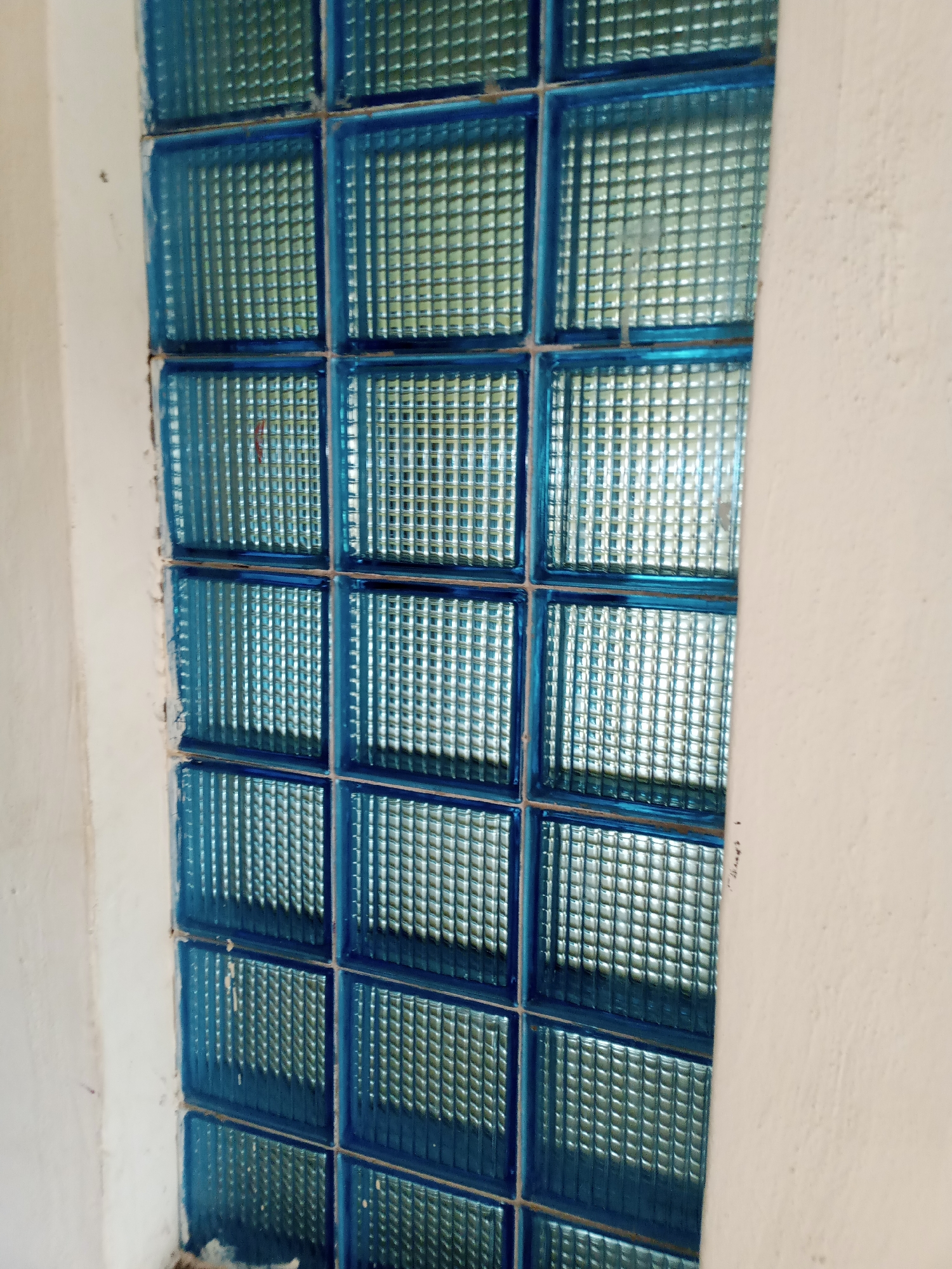 A close-up, vertical shot shows a wall made of blue glass blocks, possibly a window or partition. The blocks are arranged in a grid pattern, with five full rows visible and a partial sixth row at the bottom. Each block has a textured, grid-like surface that refracts light, creating a shimmering effect. The surrounding wall is painted a light, off-white color and appears to be made of concrete or plaster, showing some rough texture and minor imperfections. The lighting suggests an indoor or shaded outdoor environment, as there are no harsh shadows. The image does not contain any people or discernible signs of life, focusing solely on the architectural elements. The scene is generic and could be found in many urban or suburban settings. The information about the location in Jalingo, Nigeria, is not visually present in the image and must be inferred or provided externally.